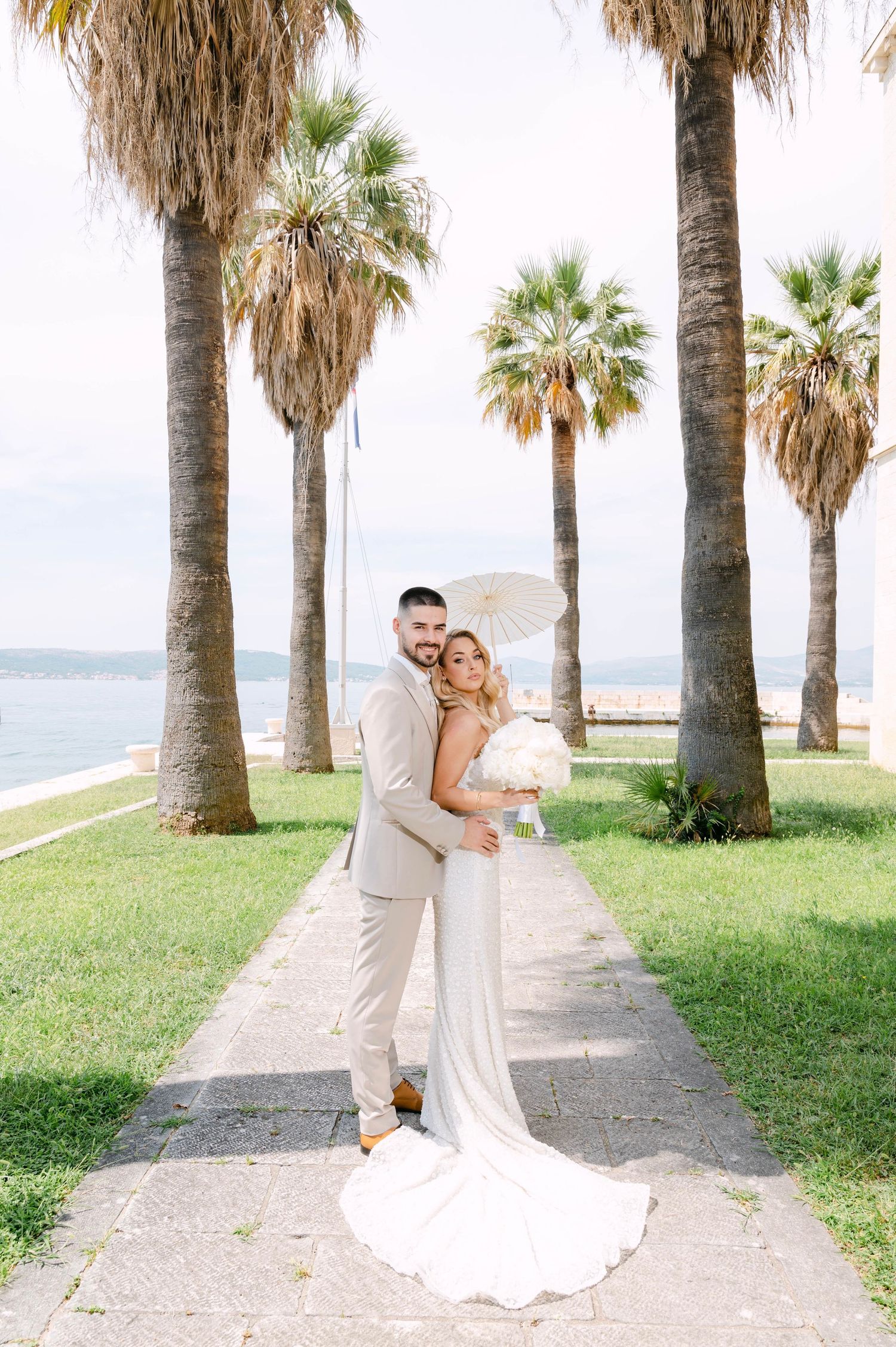 Romantic wedding couple embraces along a palm tree-lined coastal path on a sunny day.