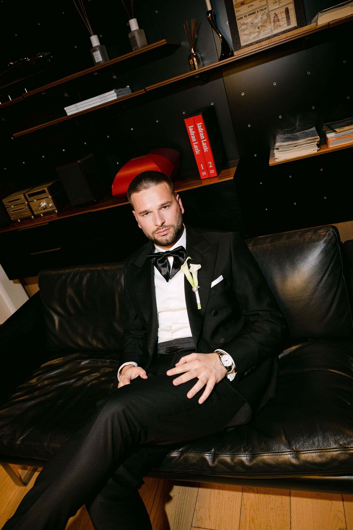 A groom in a black tuxedo with bow tie and boutonniere sits confidently on a leather sofa in a dark, elegant room.
