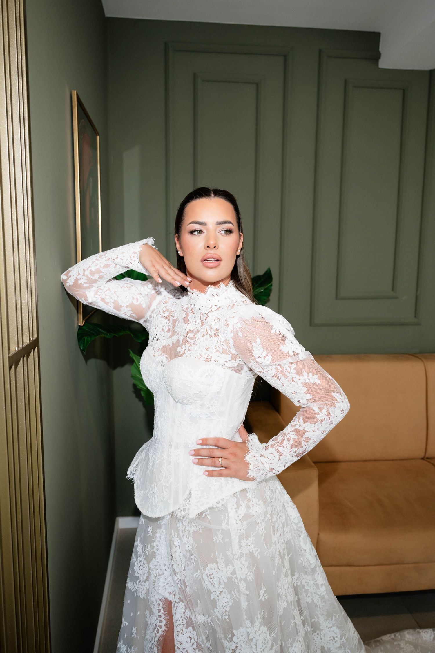 Elegant bride in white lace long-sleeve wedding gown with corset waist posing in a green-walled luxury interior.