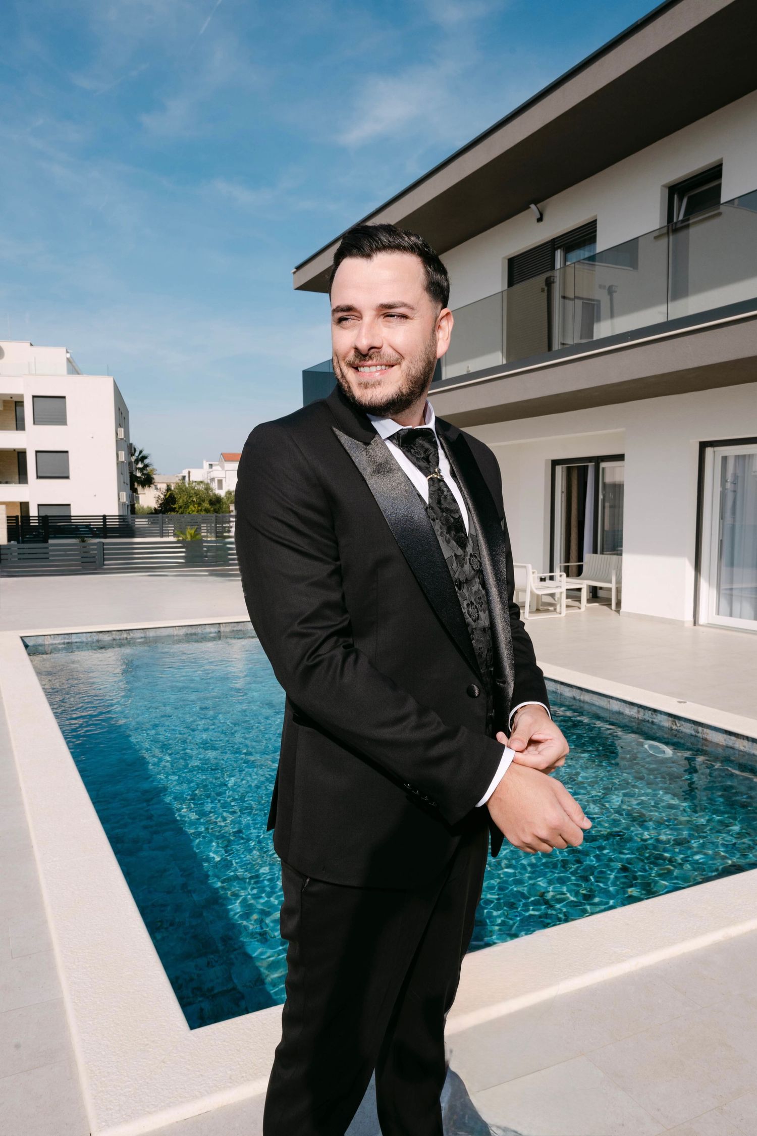 A person in a black suit and tie stands beside a modern swimming pool with a luxury house in the background.