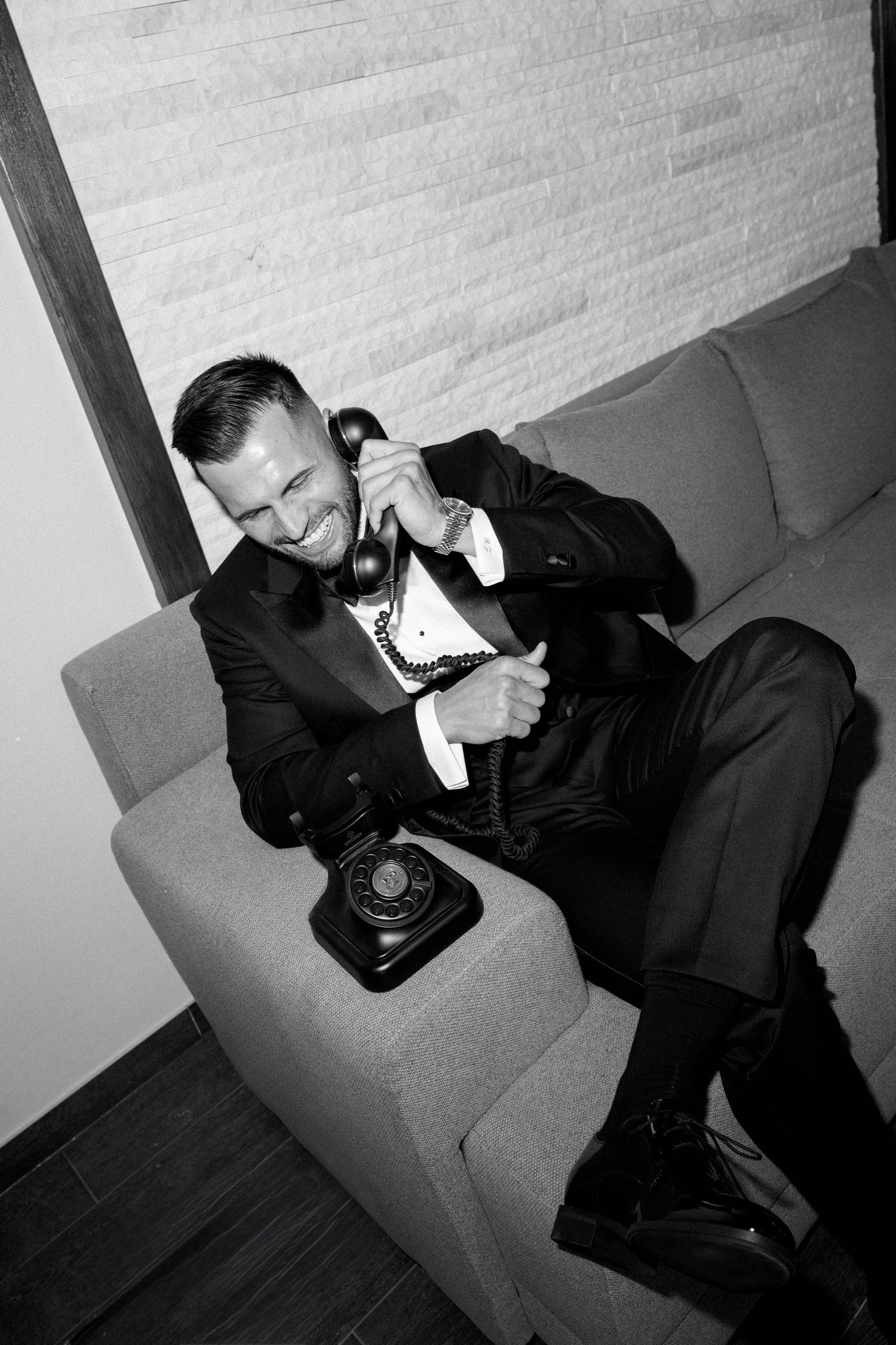 Man in tuxedo smiling while talking on vintage rotary phone, seated on sofa in black and white photo.