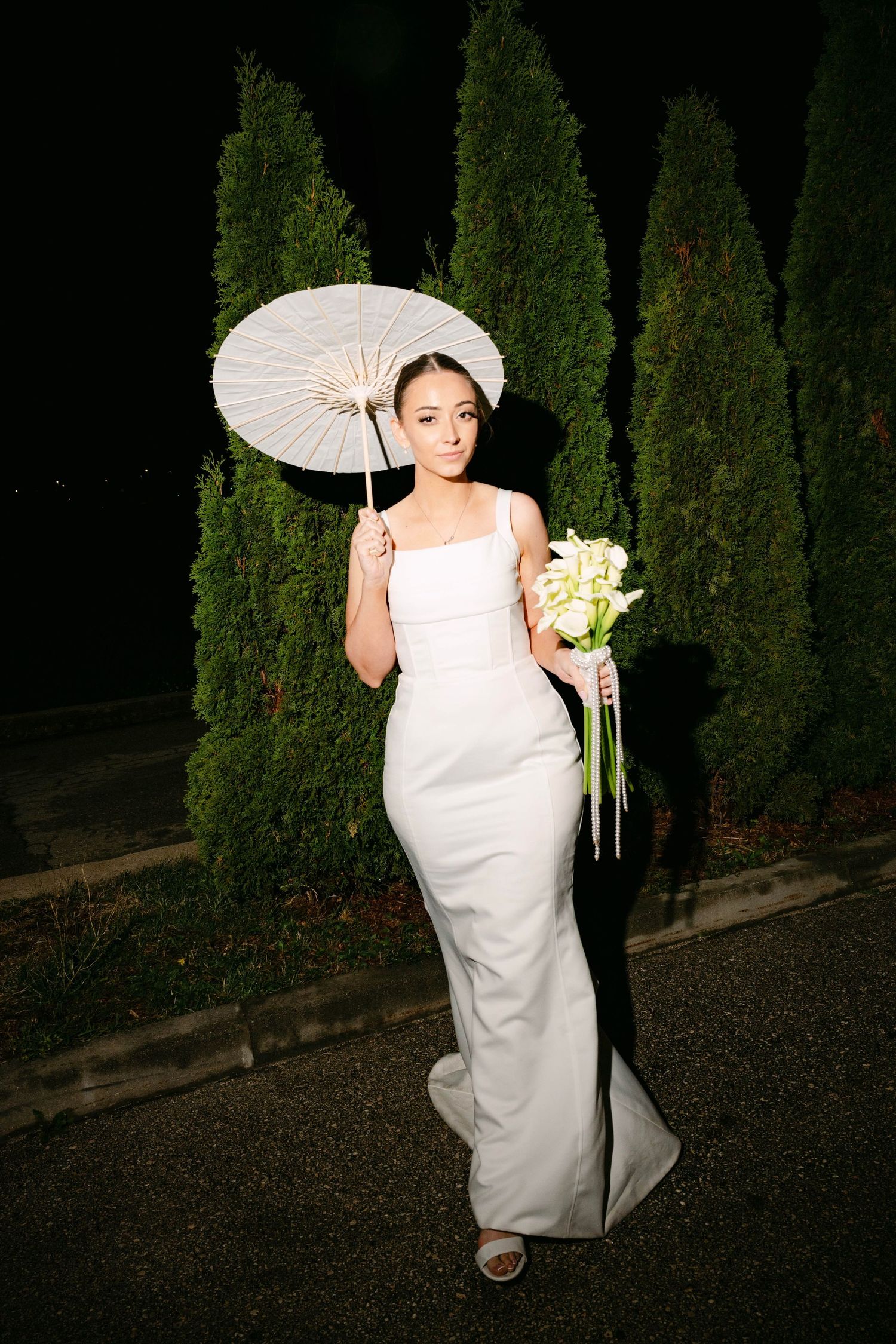Elegant figure in white wedding gown holds parasol and bouquet while standing against backdrop of tall green cypress trees.