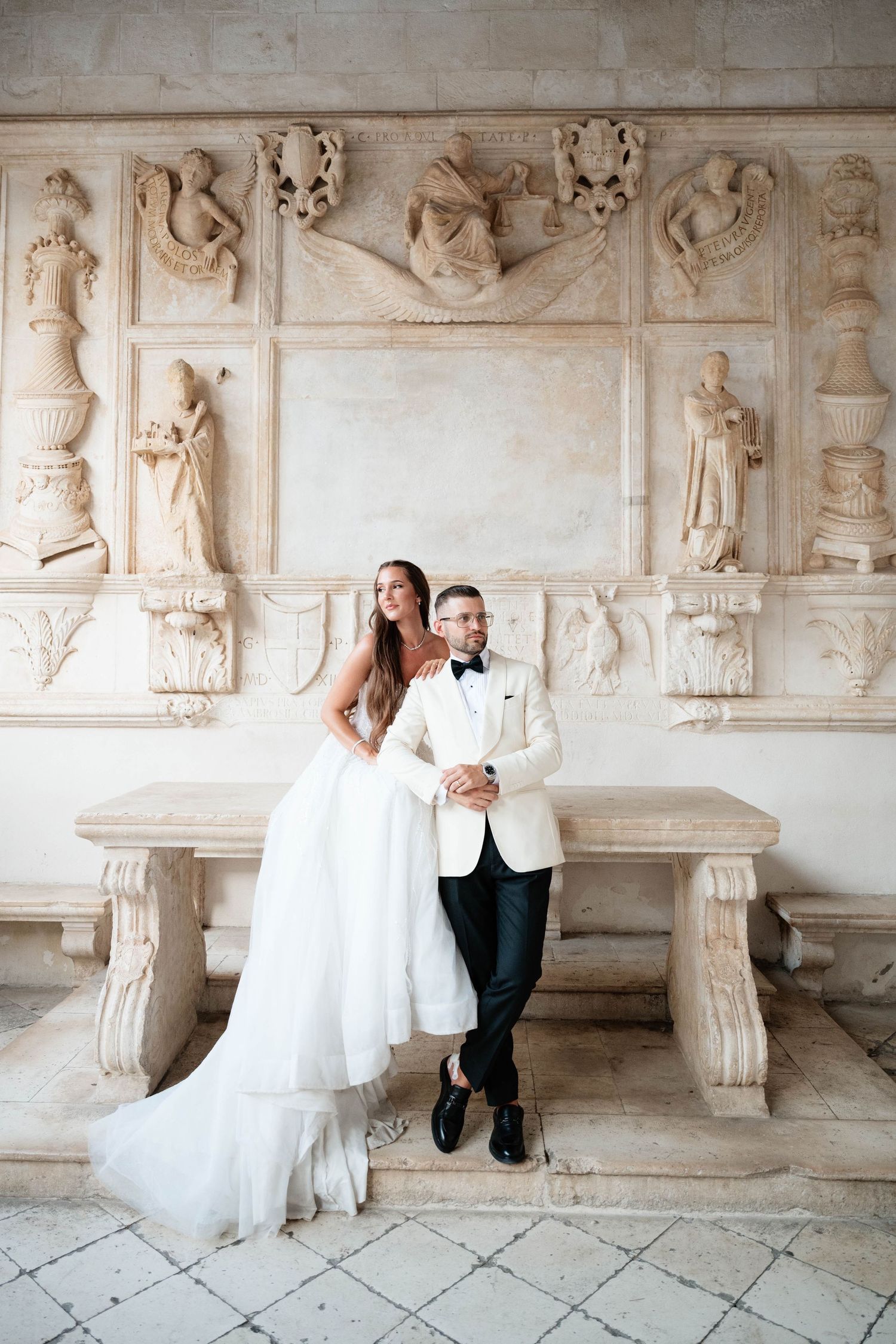 Bride in white gown and groom in ivory tuxedo pose before ornate marble relief sculptures at elegant venue.