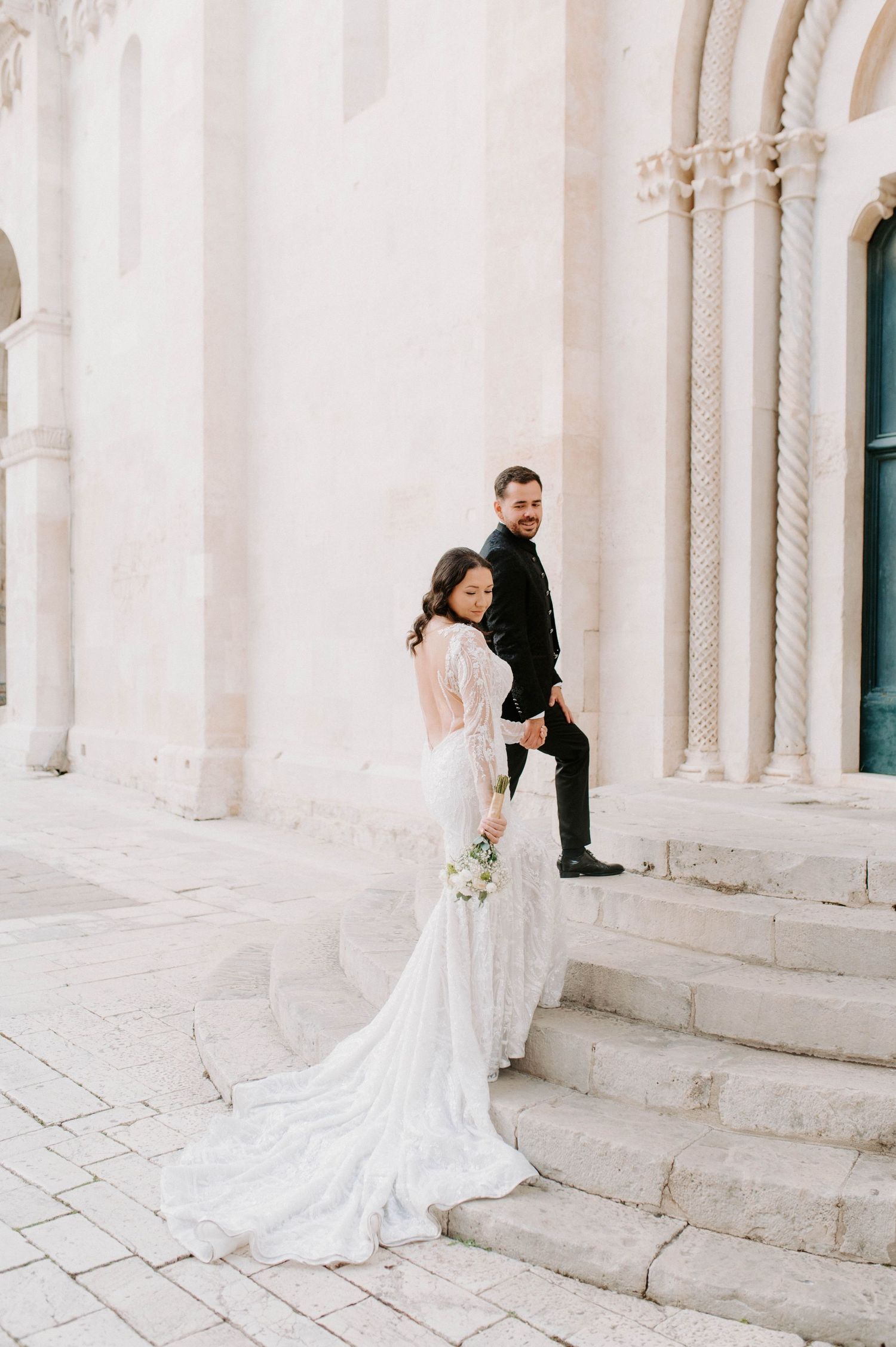 Elegant wedding couple poses on stone church steps with flowing white dress and classic architecture in background.