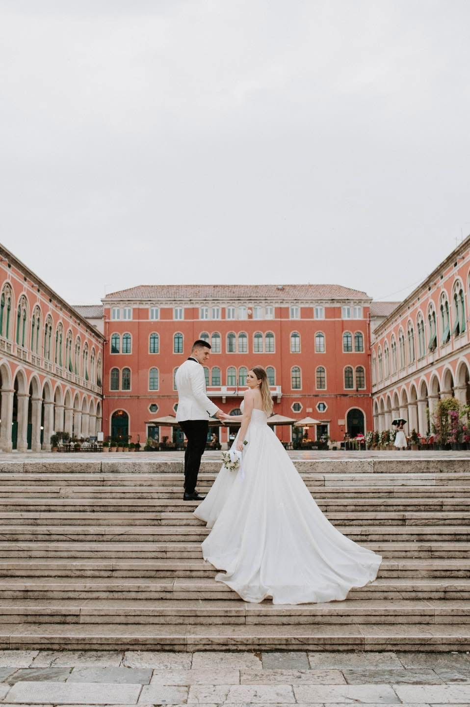 Bride and groom stand on grand red palazzo steps in romantic Italian square.