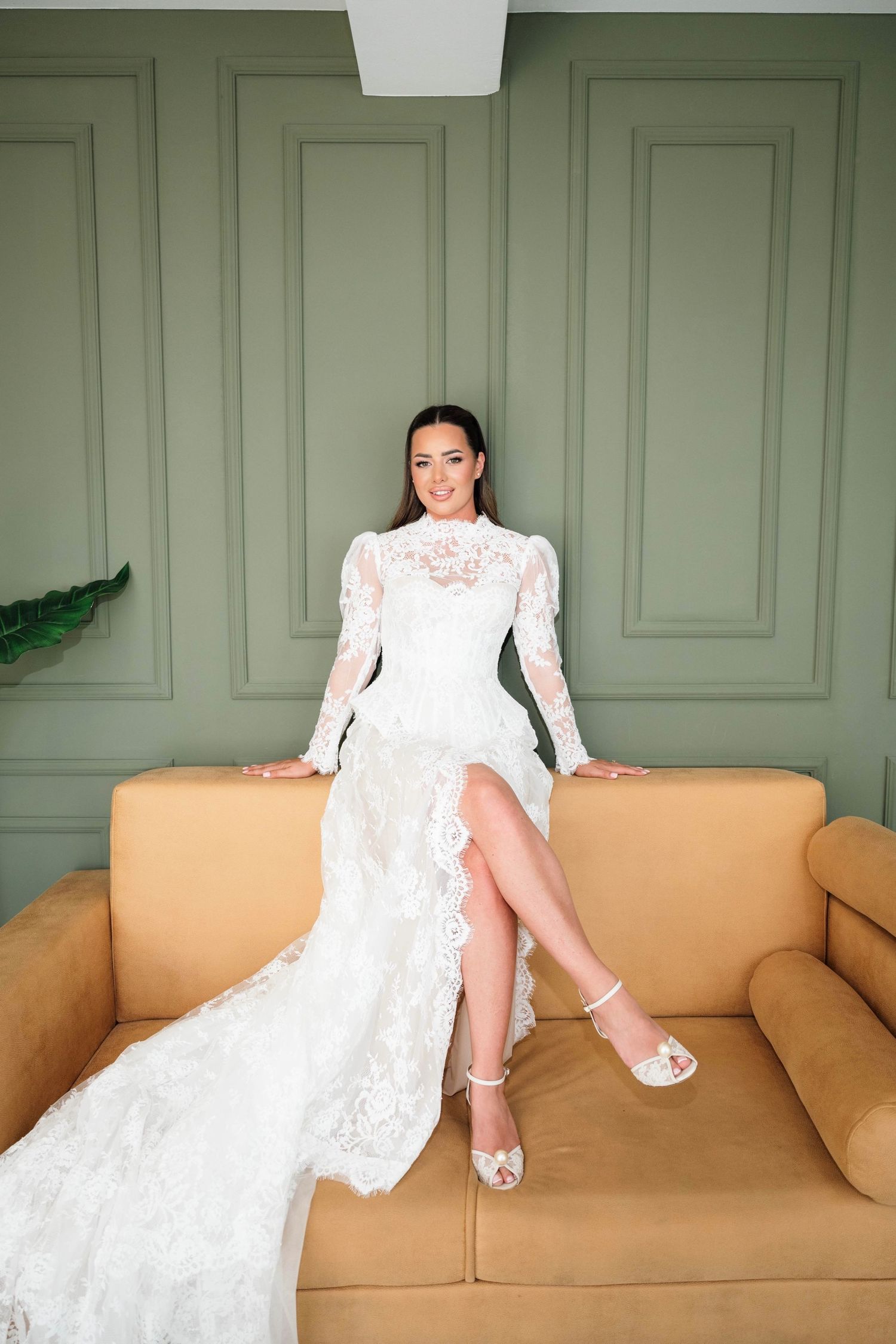 Smiling bride in long-sleeve lace wedding gown with thigh-high slit sits on tan sofa against sage green wall.