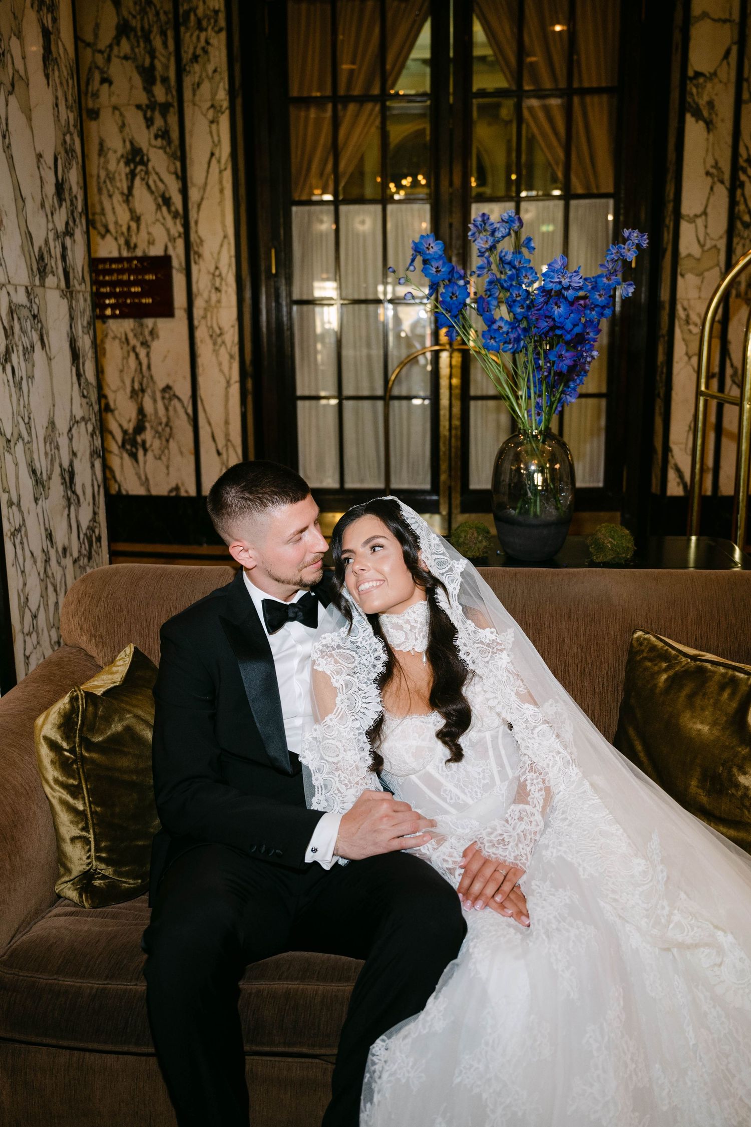 Bride in lace gown and veil sits with tuxedo-clad groom on velvet sofa, blue floral arrangement behind them.