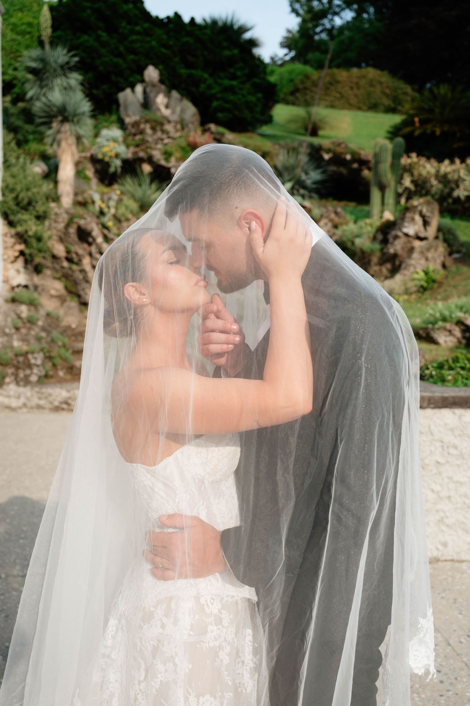 Bride and groom sharing a romantic moment under a wedding veil in a lush garden setting.