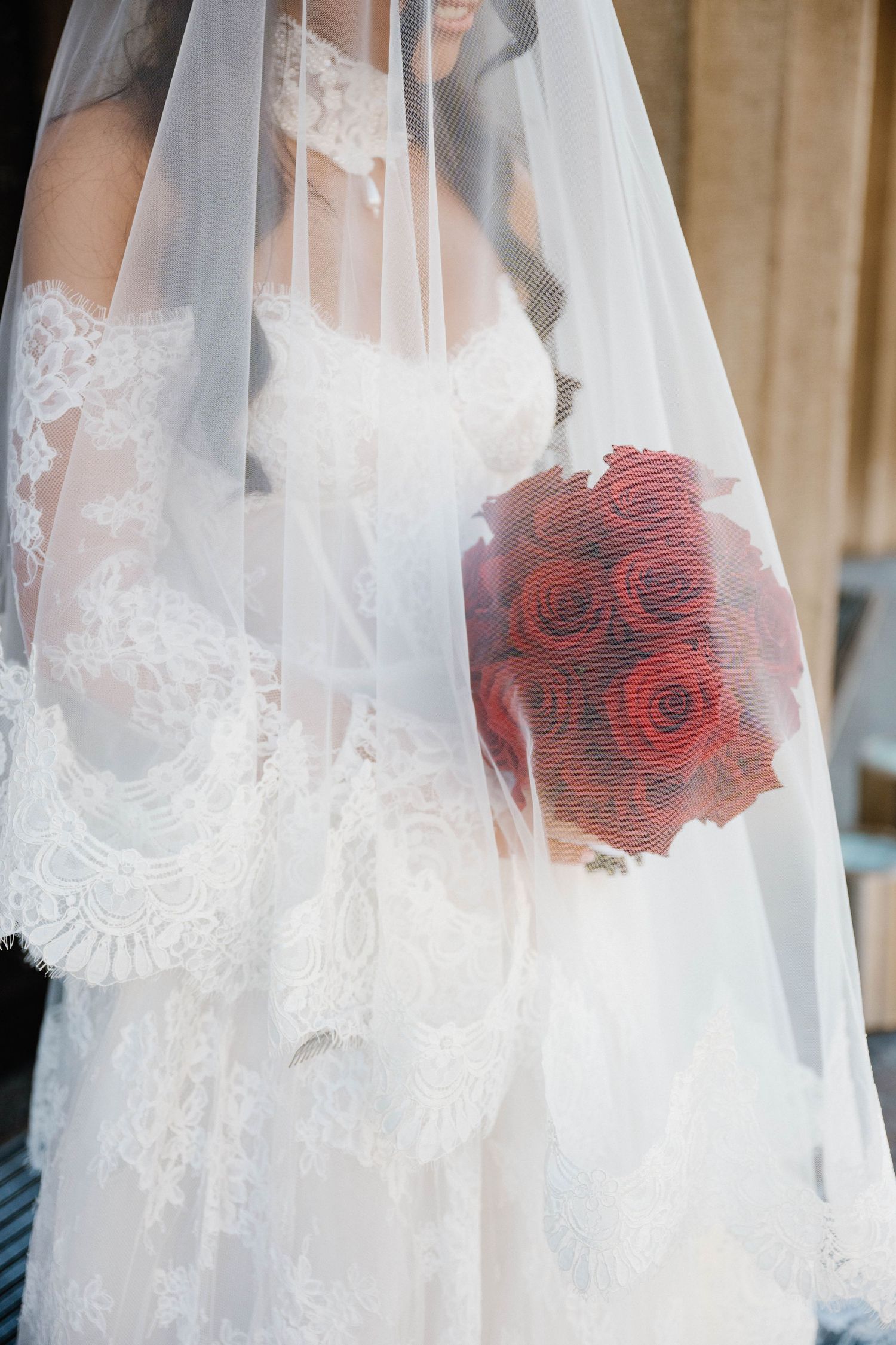 Bride in lace wedding dress holding red rose bouquet under flowing white veil.