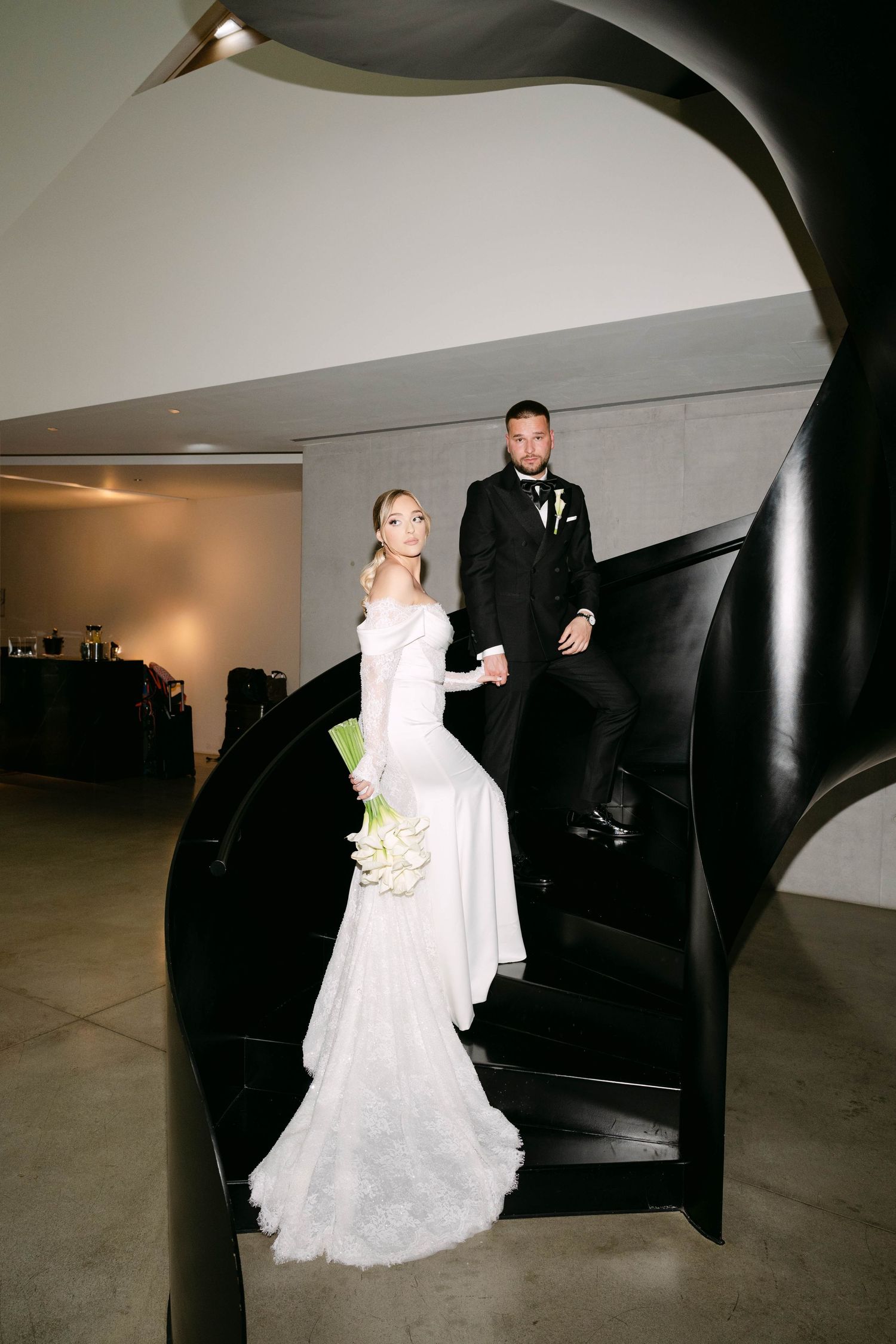 Bride in white mermaid gown and groom in black tuxedo pose on dramatic black spiral staircase at wedding venue.