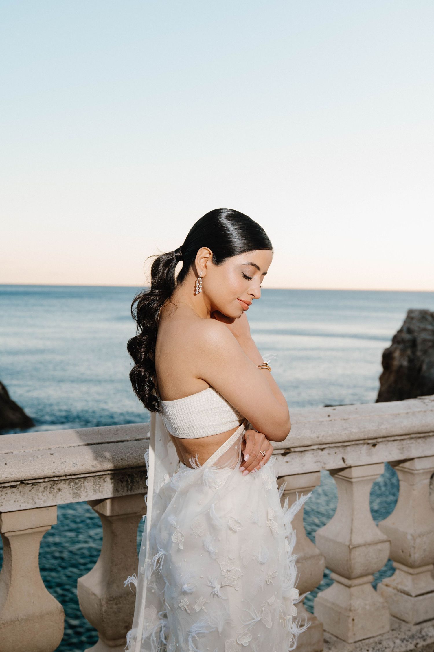 Bride in a strapless white dress poses by ornate stone balcony overlooking ocean at sunset.