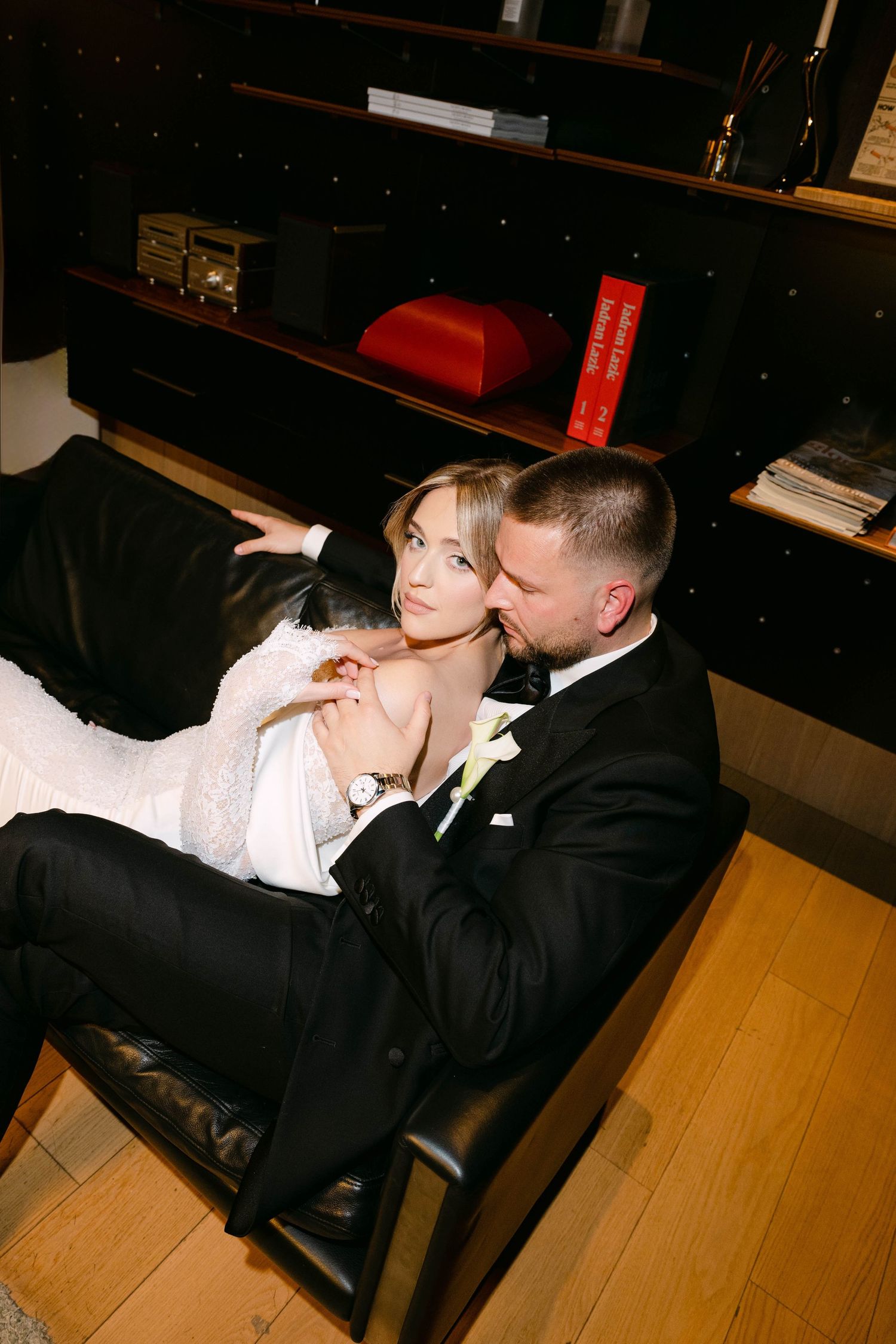 A bride and groom in wedding attire share an intimate moment seated together on a leather chair near a bookshelf.