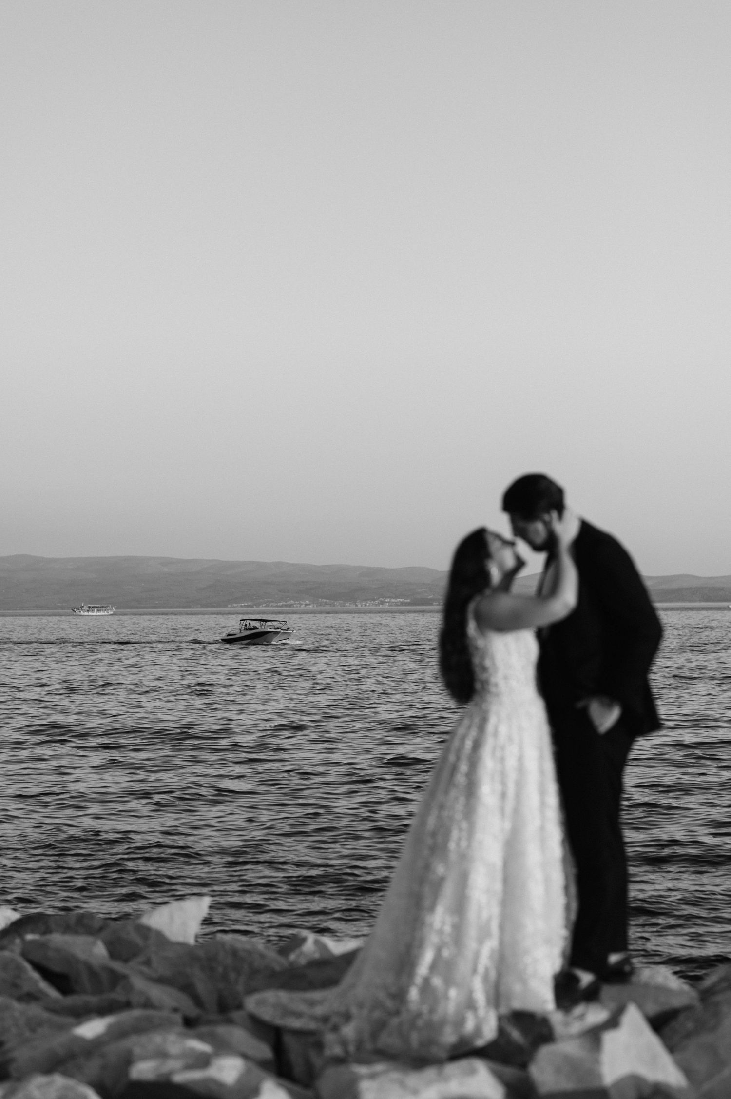A romantic black and white wedding photo of a couple embracing on rocky shore with ocean and misty horizon in background.