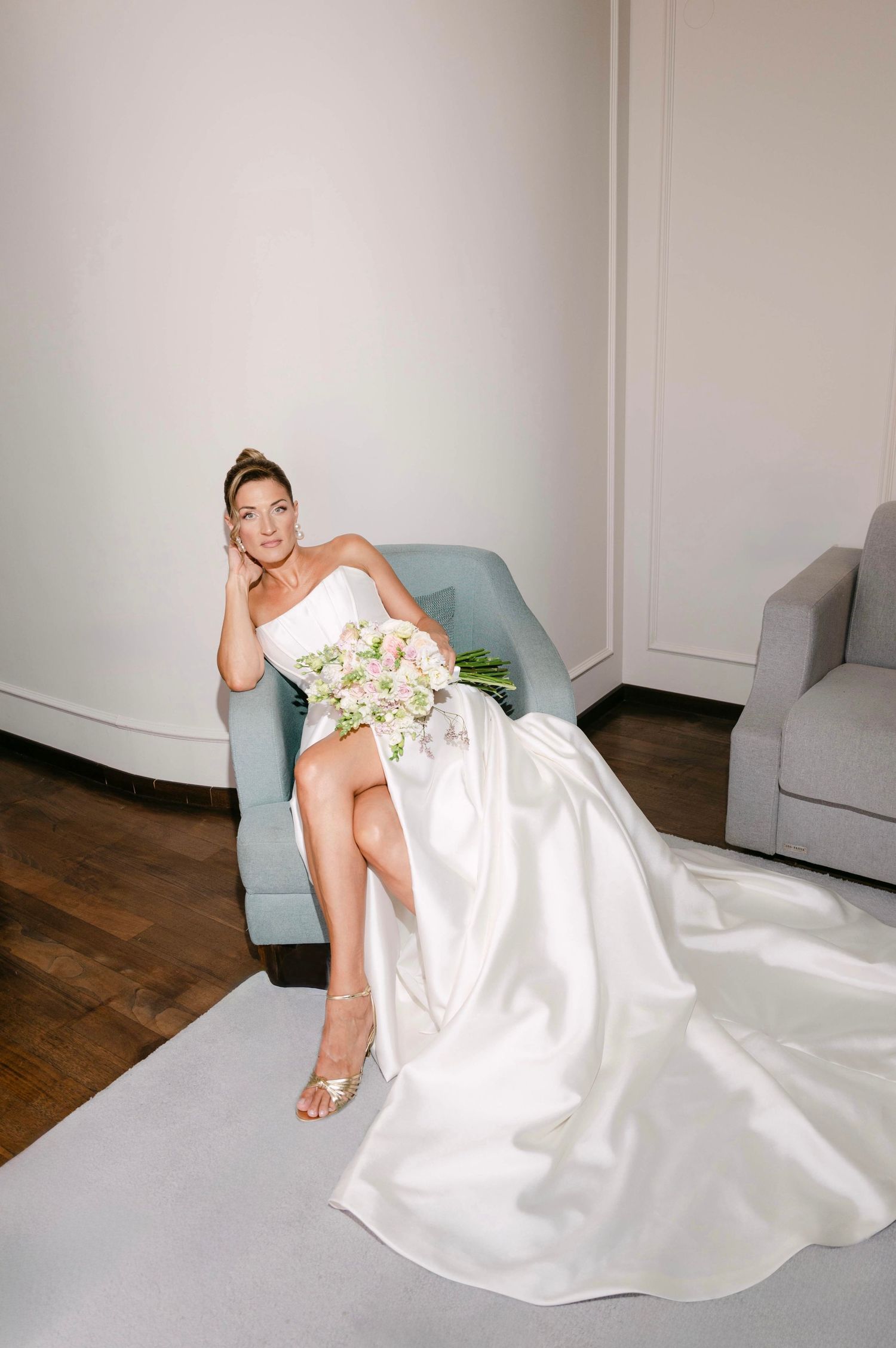 A bride in a white strapless gown reclines elegantly on a blue chair holding a bouquet of flowers.