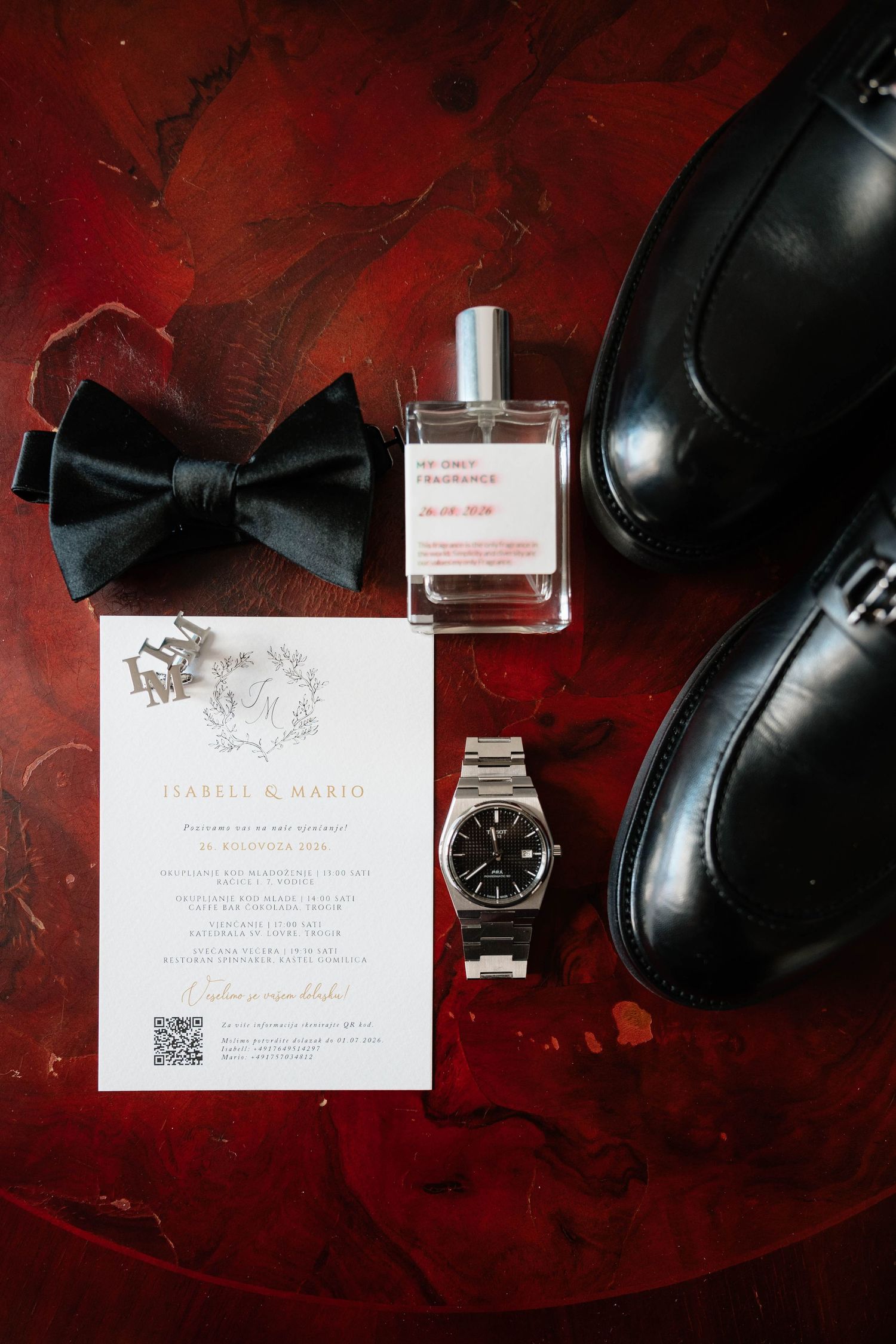 Groom wedding flat lay with black bow tie, dress shoes, watch, cologne, cufflinks, and elegant invitation on red surface.