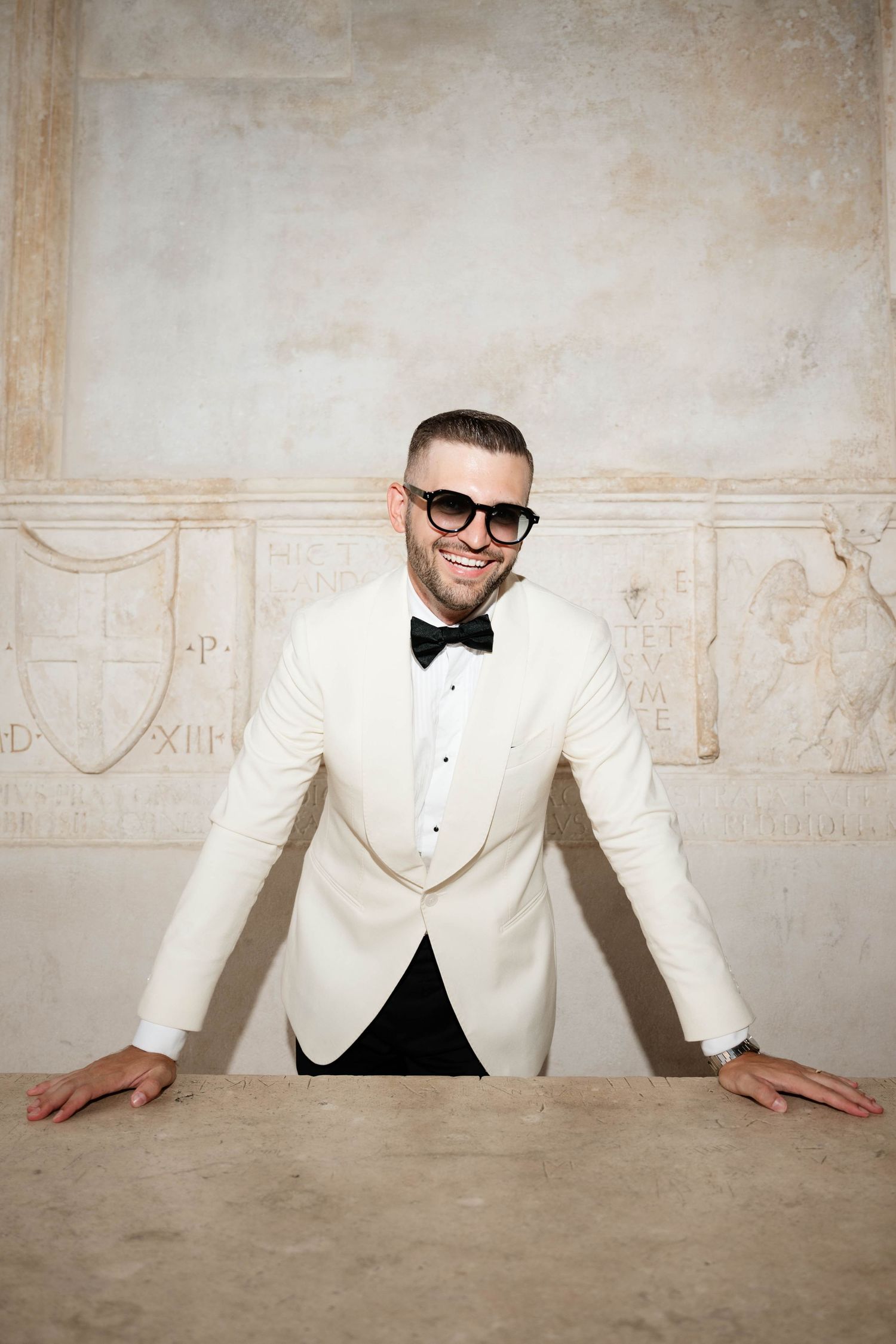 Smiling man in white tuxedo jacket and black bow tie leans on stone table in elegant vintage interior.