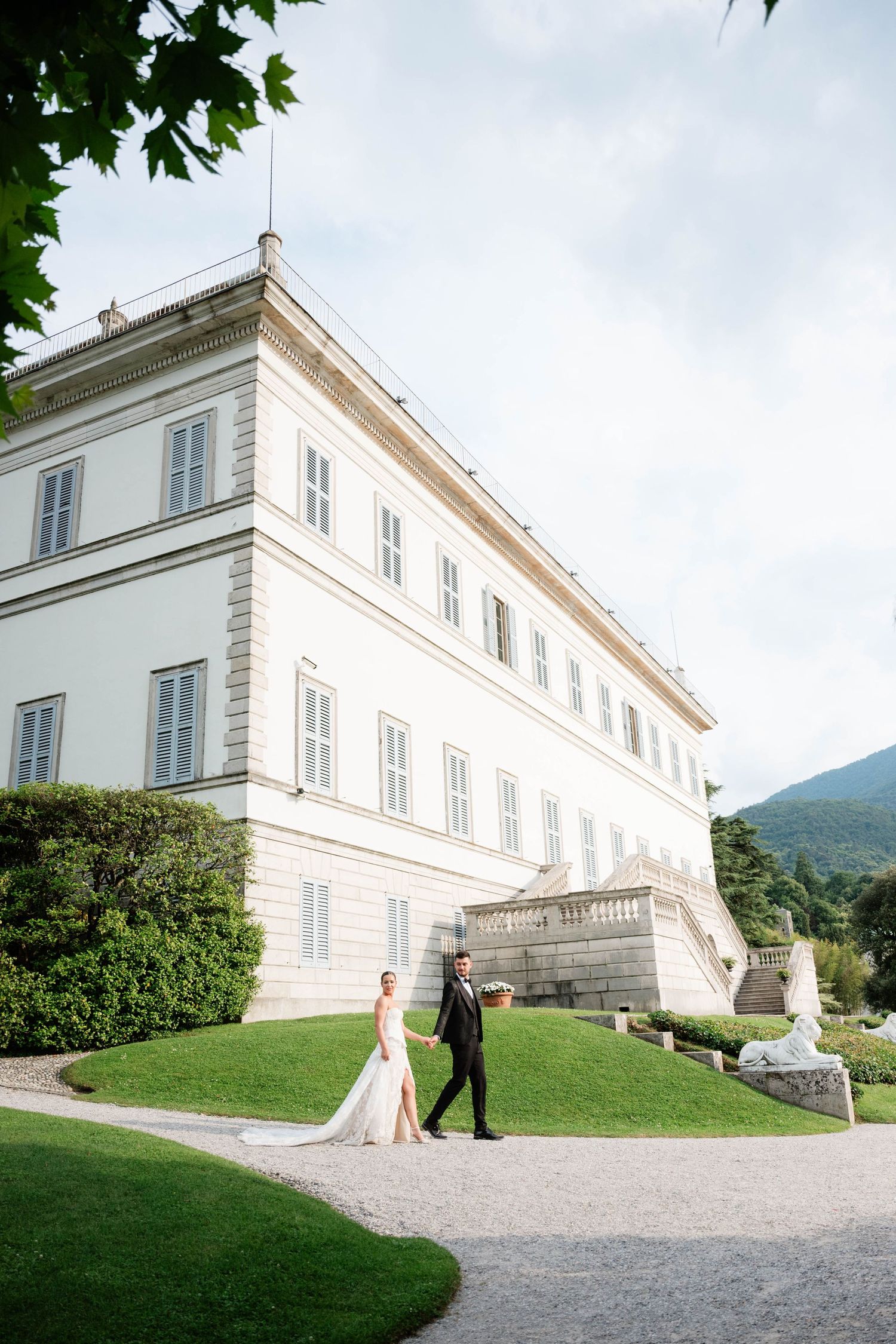 Bride and groom walking on garden path beside elegant white Italian villa on Lake Como wedding day.