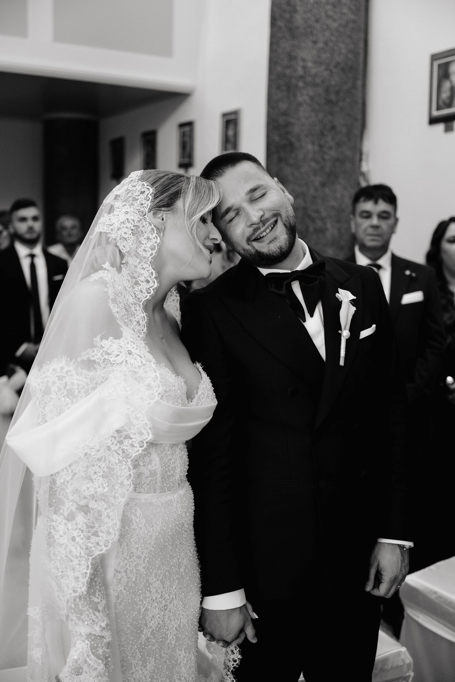 Black and white photo of a bride kissing the groom on the cheek as he smiles joyfully during their wedding ceremony.