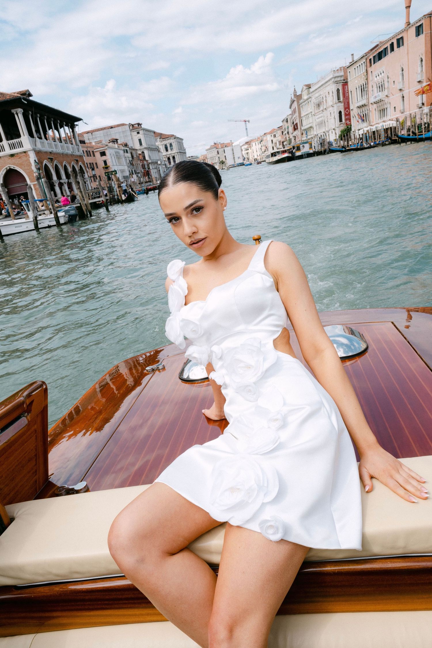A person in a white ruffle dress poses on a wooden boat along Venice's Grand Canal with historic buildings in the background.
