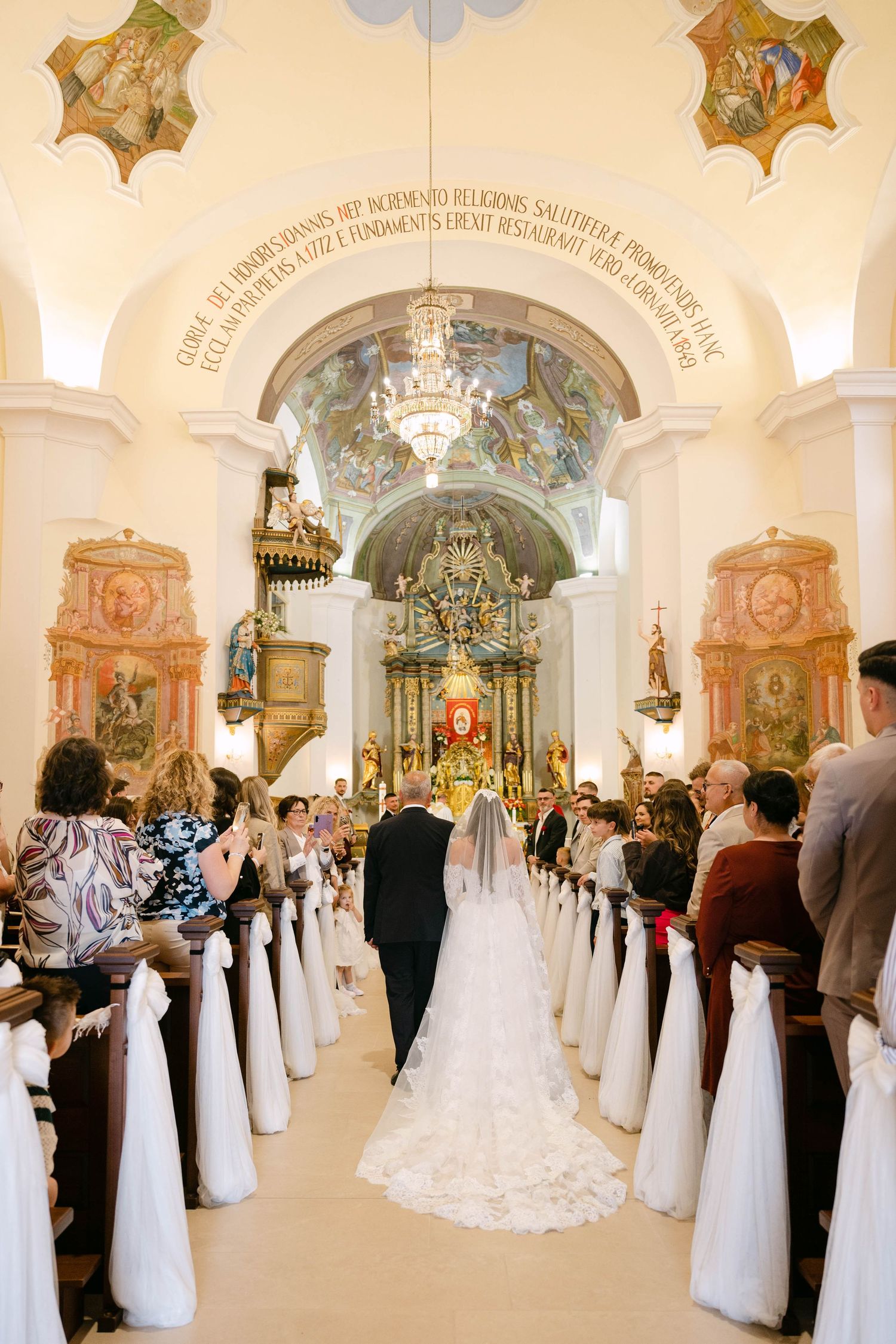 Bride walks down the aisle of an ornate church with vaulted ceilings and gilded altar during a wedding ceremony.