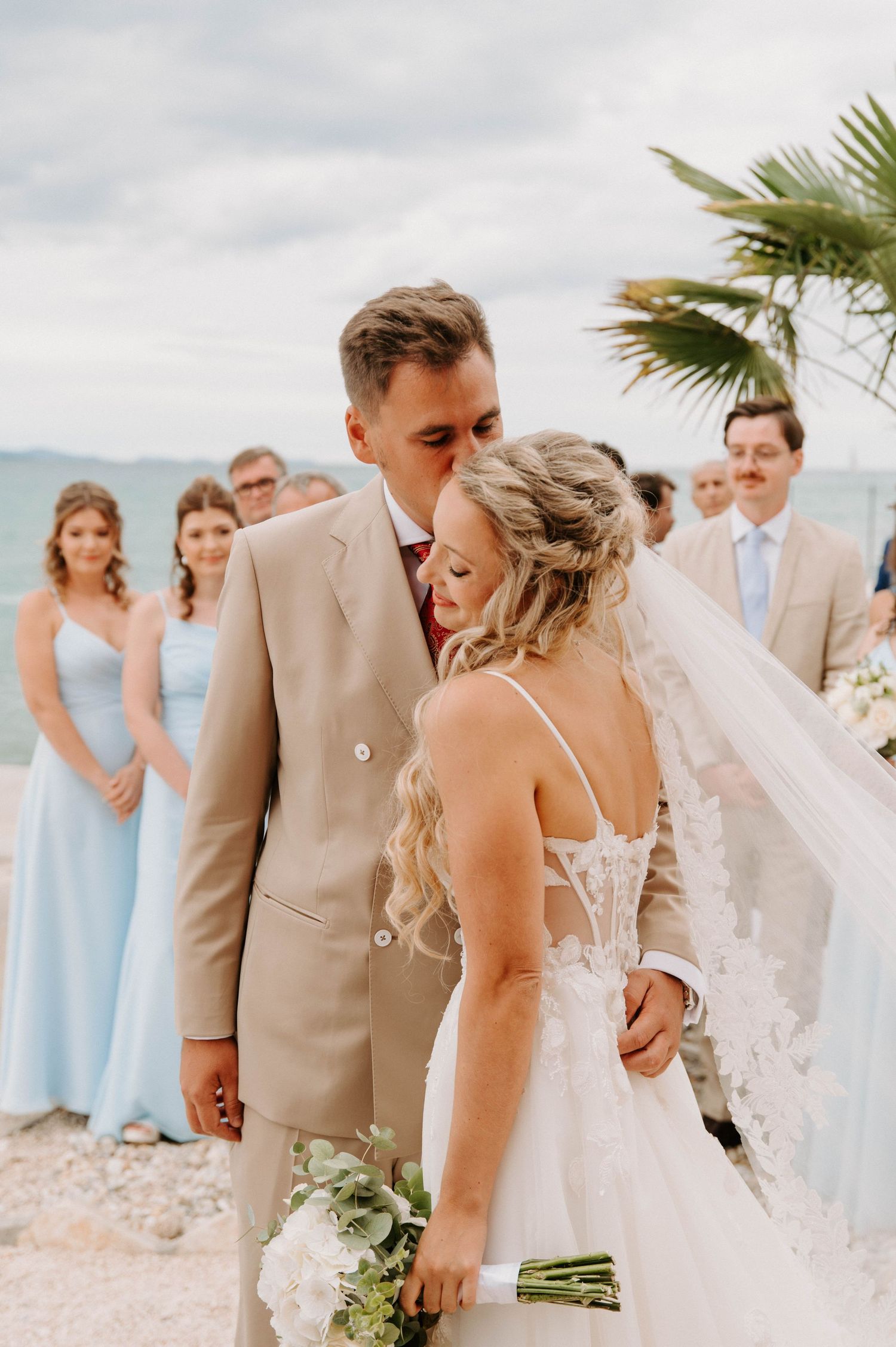Beachside wedding scene with couple in light formal attire and bridal party in pastel blue dresses.