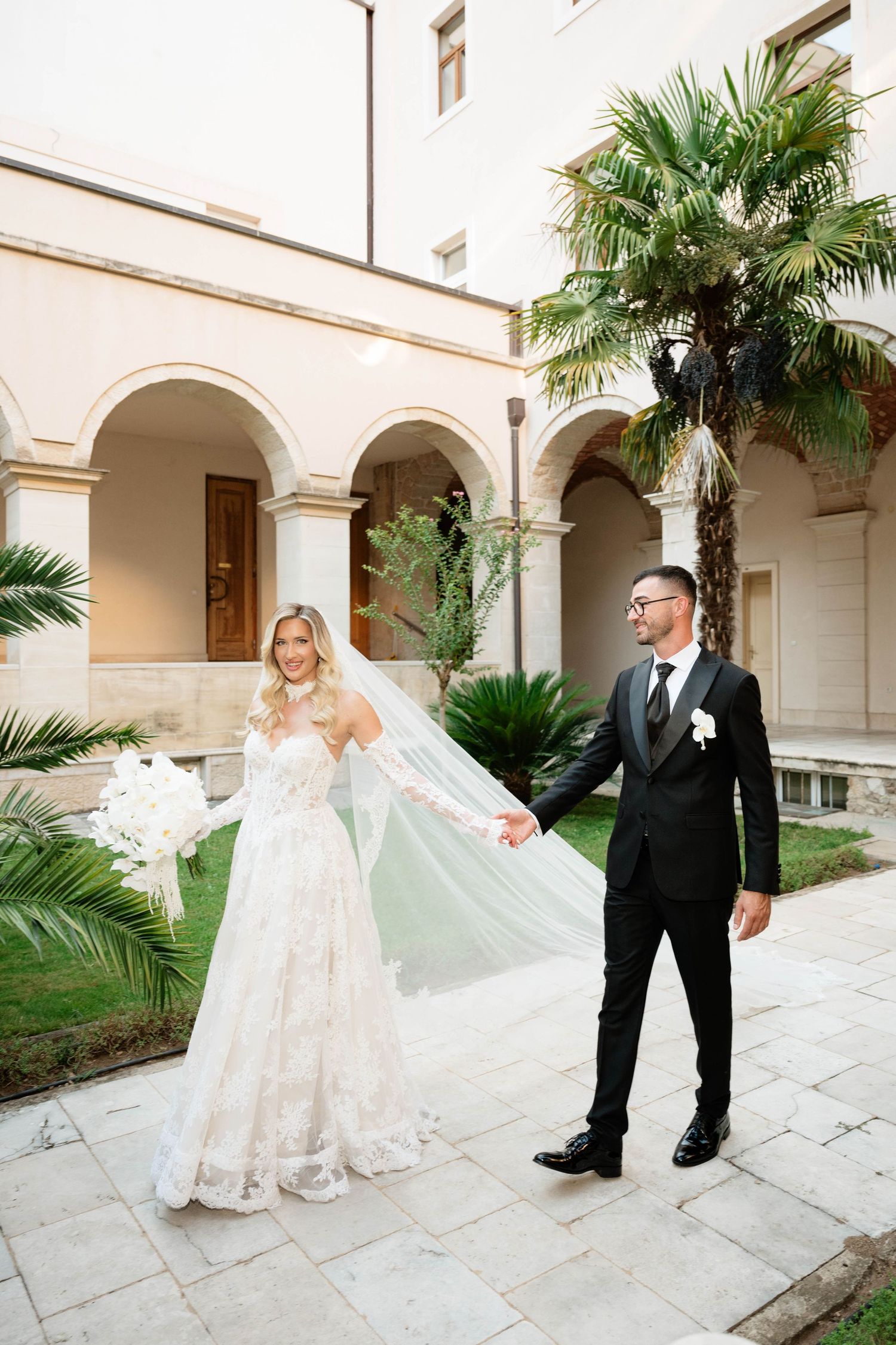 Bride in lace gown and groom in black tuxedo hold hands in elegant courtyard with palm trees.