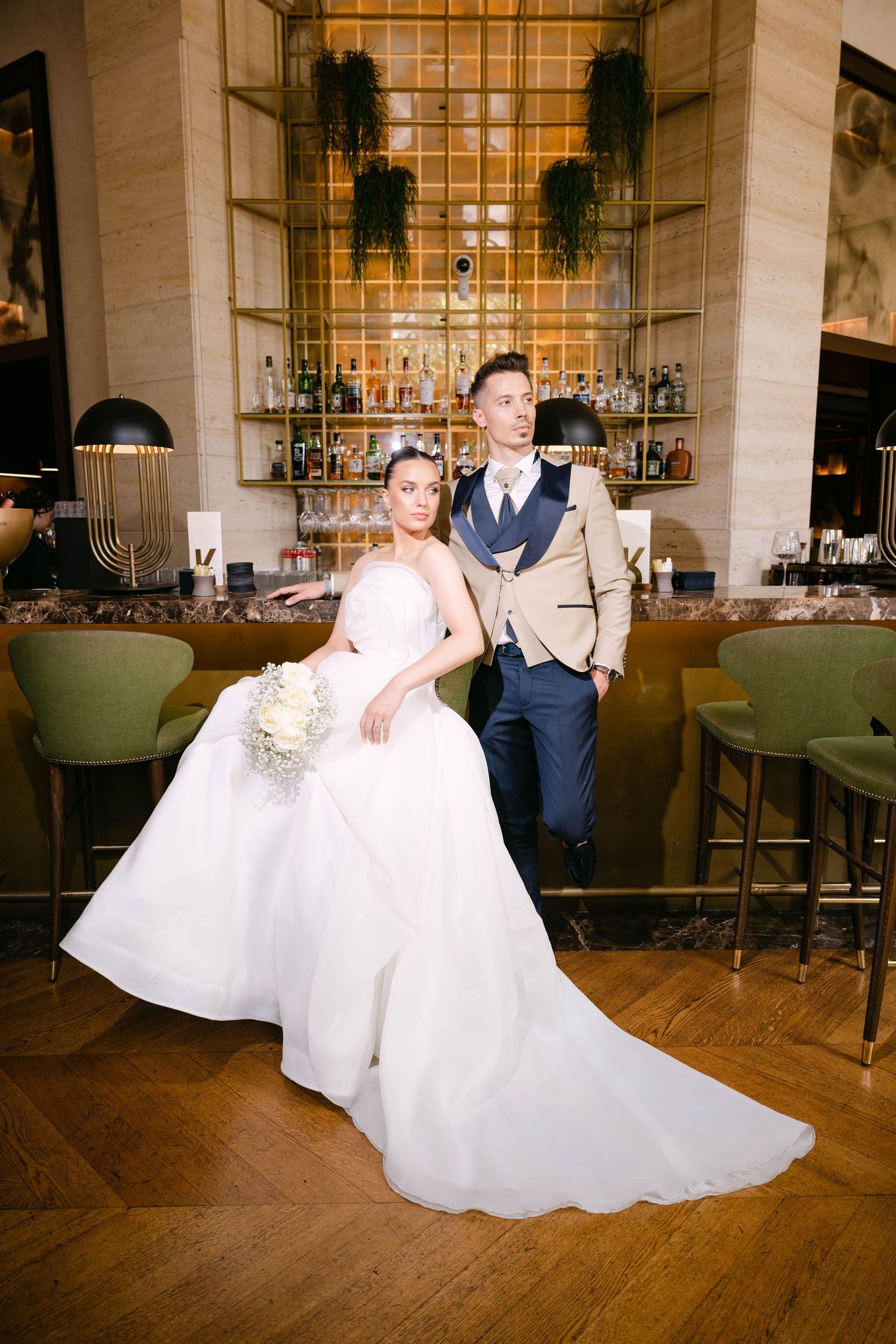 Elegant wedding couple poses in upscale restaurant bar with wood floors and decorative greenery on display shelves.