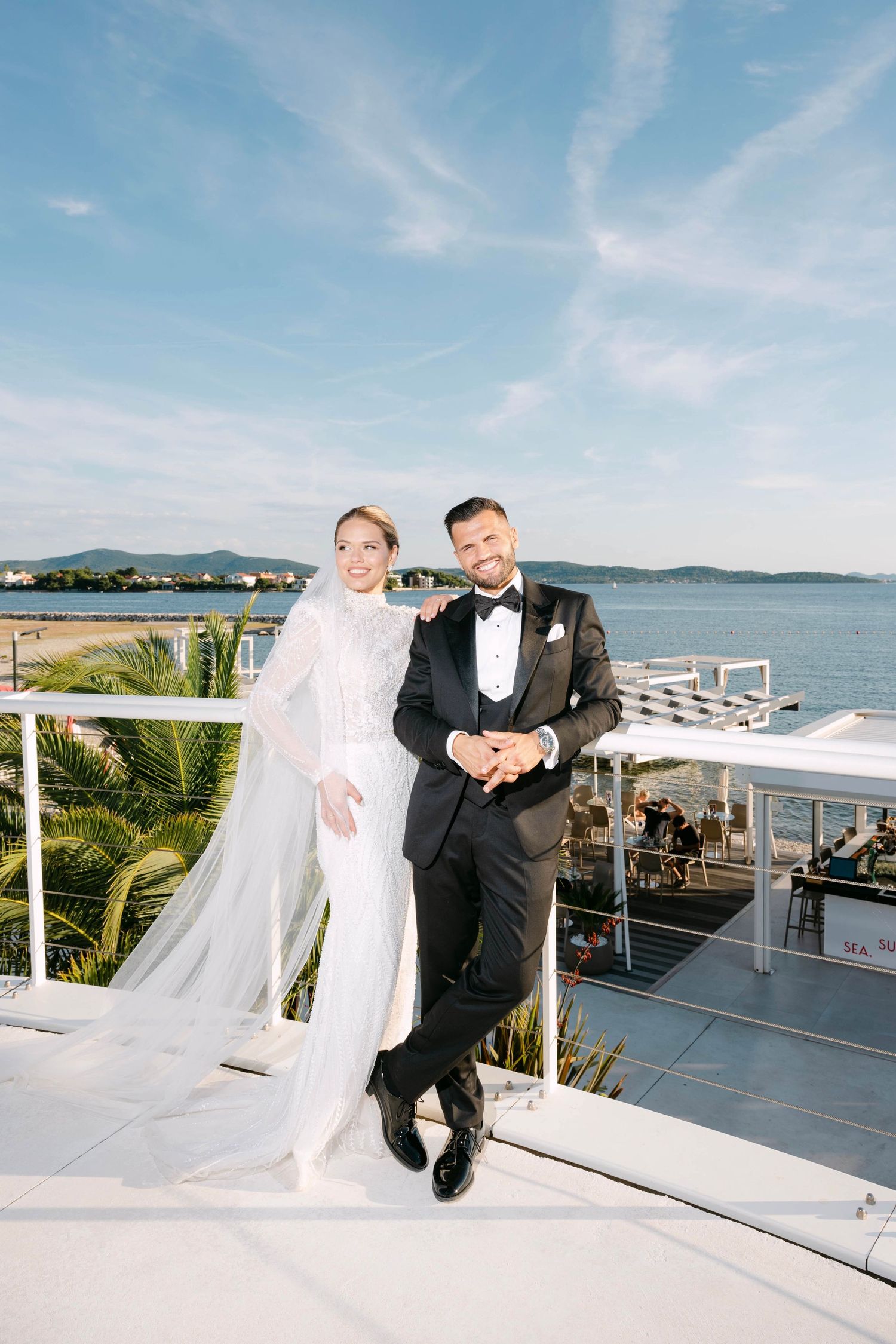 Bride in white gown with veil and groom in black tuxedo posing on rooftop terrace with sea view backdrop.