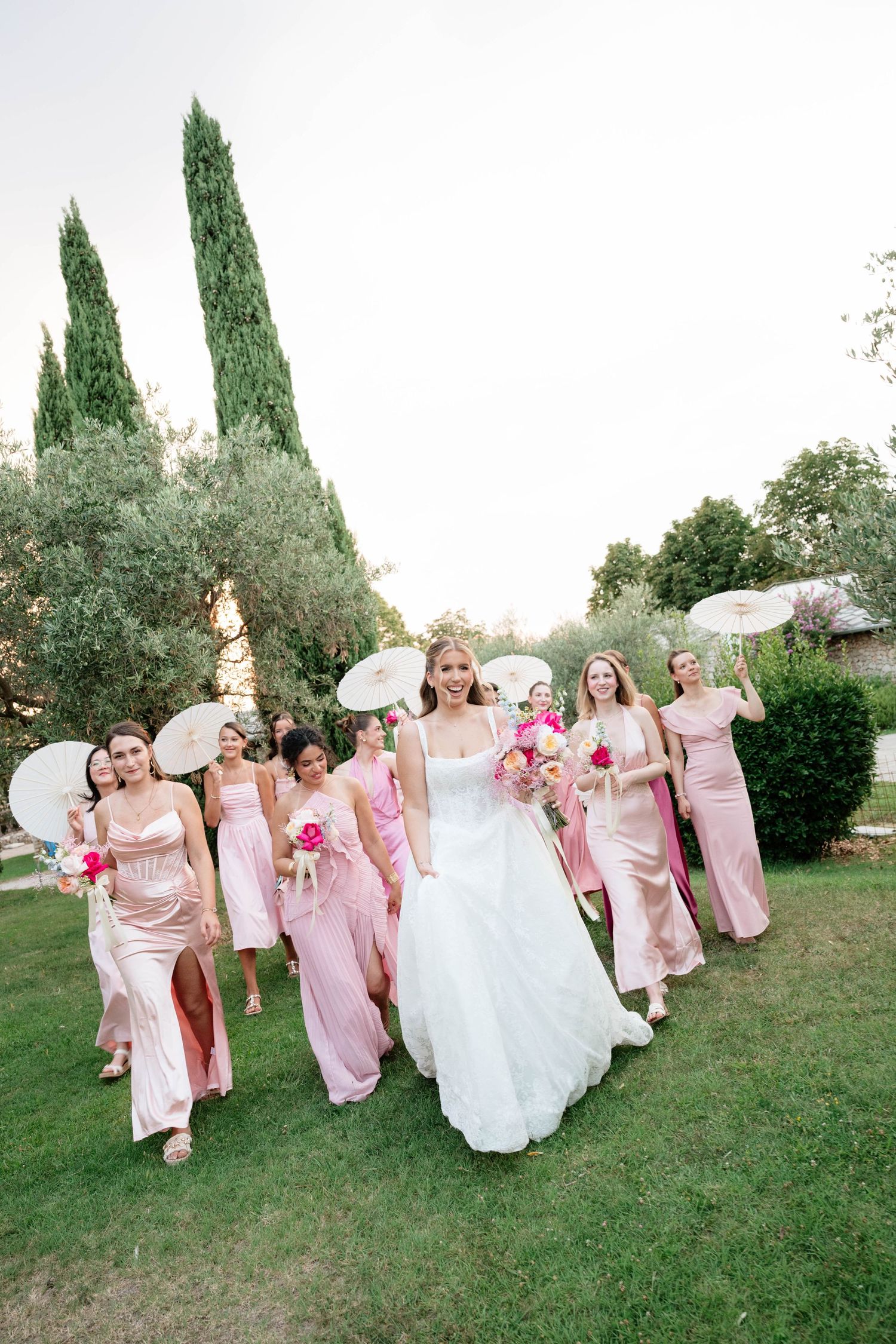 Bride in white gown walks with bridesmaids in pink dresses holding parasols and bouquets in a lush garden.