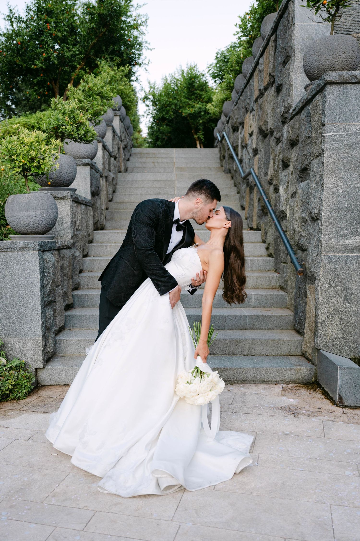 A romantic wedding photo on stone steps with a bride in white gown holding flowers and groom in black suit sharing a kiss.