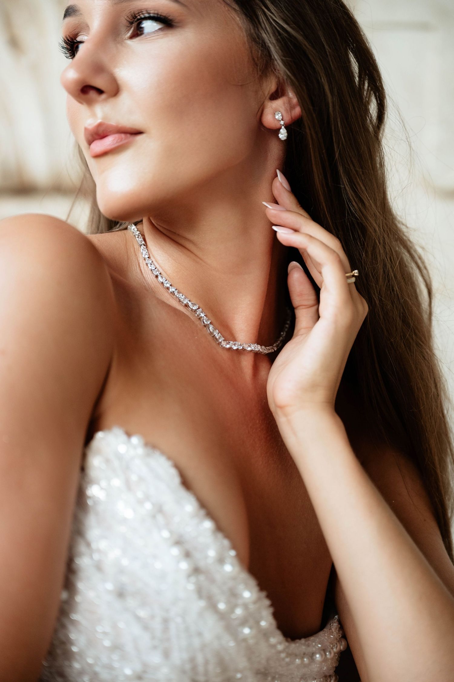Bride in white gown wearing diamond necklace and earrings, touching her ear elegantly.