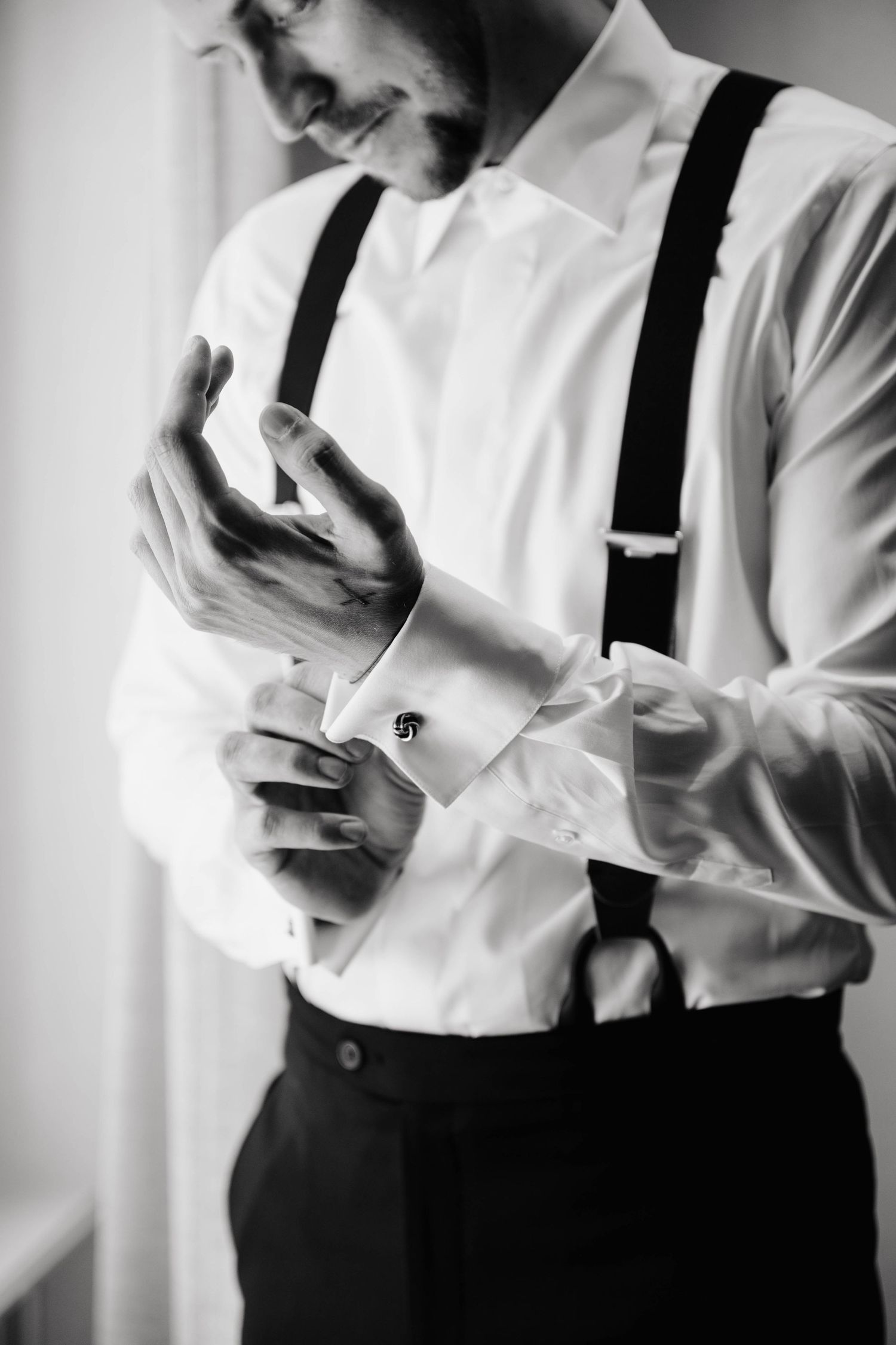 Black and white photo of a groom fastening cufflinks on a white dress shirt with suspenders, smiling while getting ready.