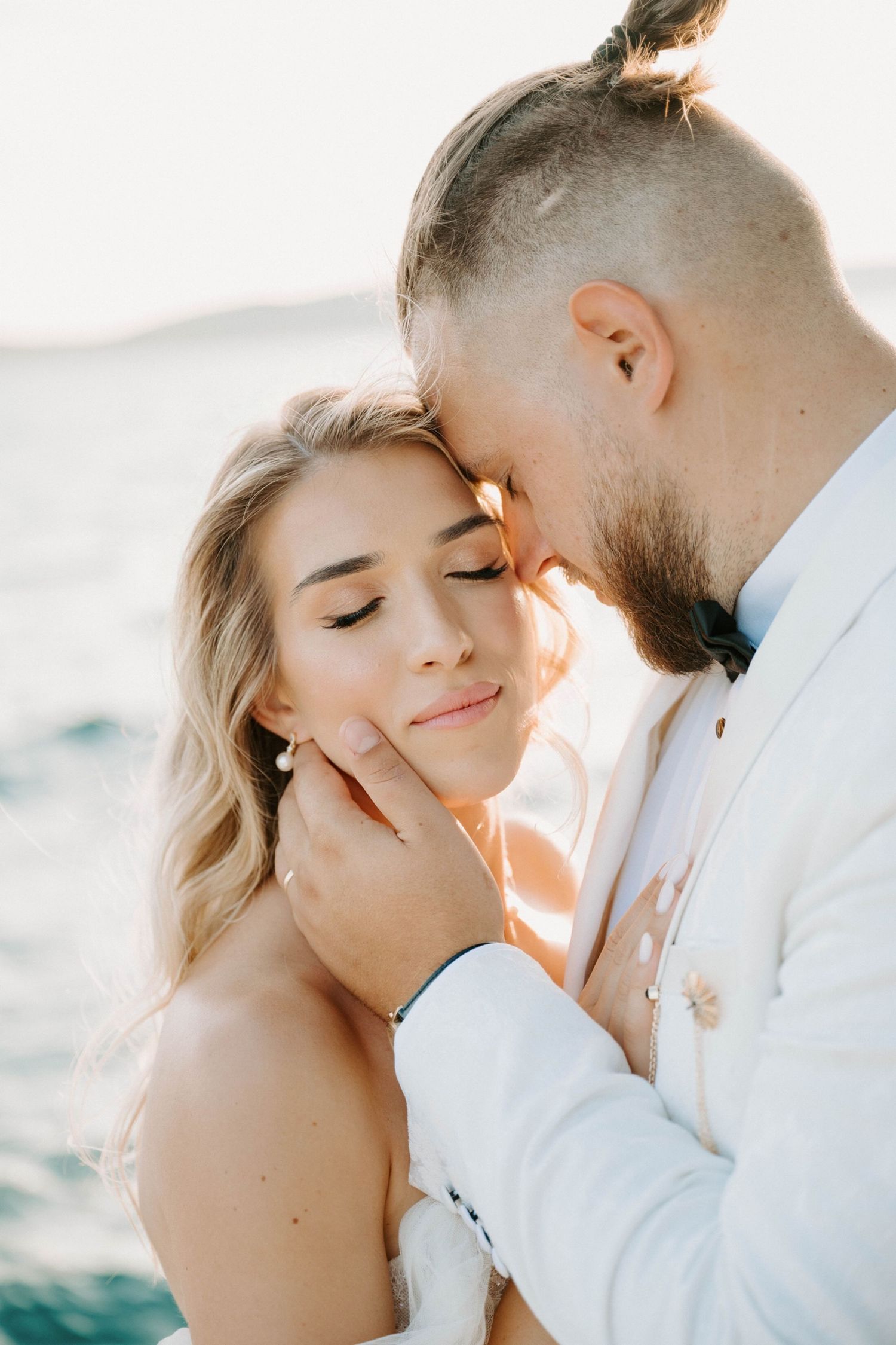 Romantic wedding couple embraces in soft sunlight by the ocean during golden hour sunset.