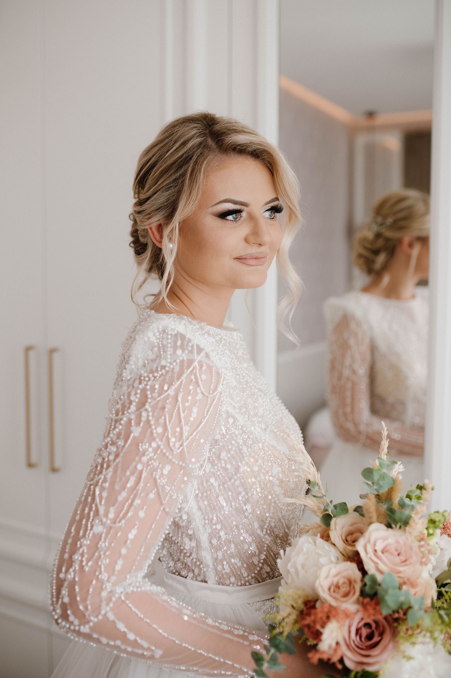 Bride wearing sequined long-sleeve gown holds peach and blush rose bouquet.