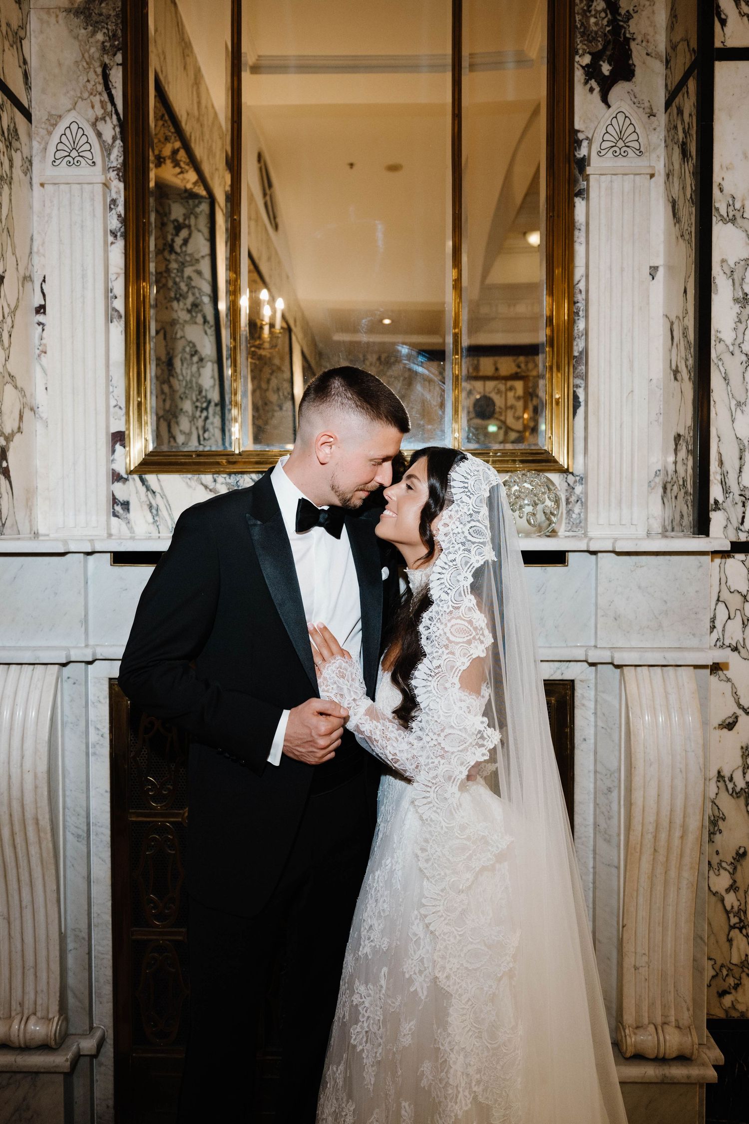 Bride and groom share a romantic moment in an elegant marble venue, she in lace gown with veil, he in black tuxedo.