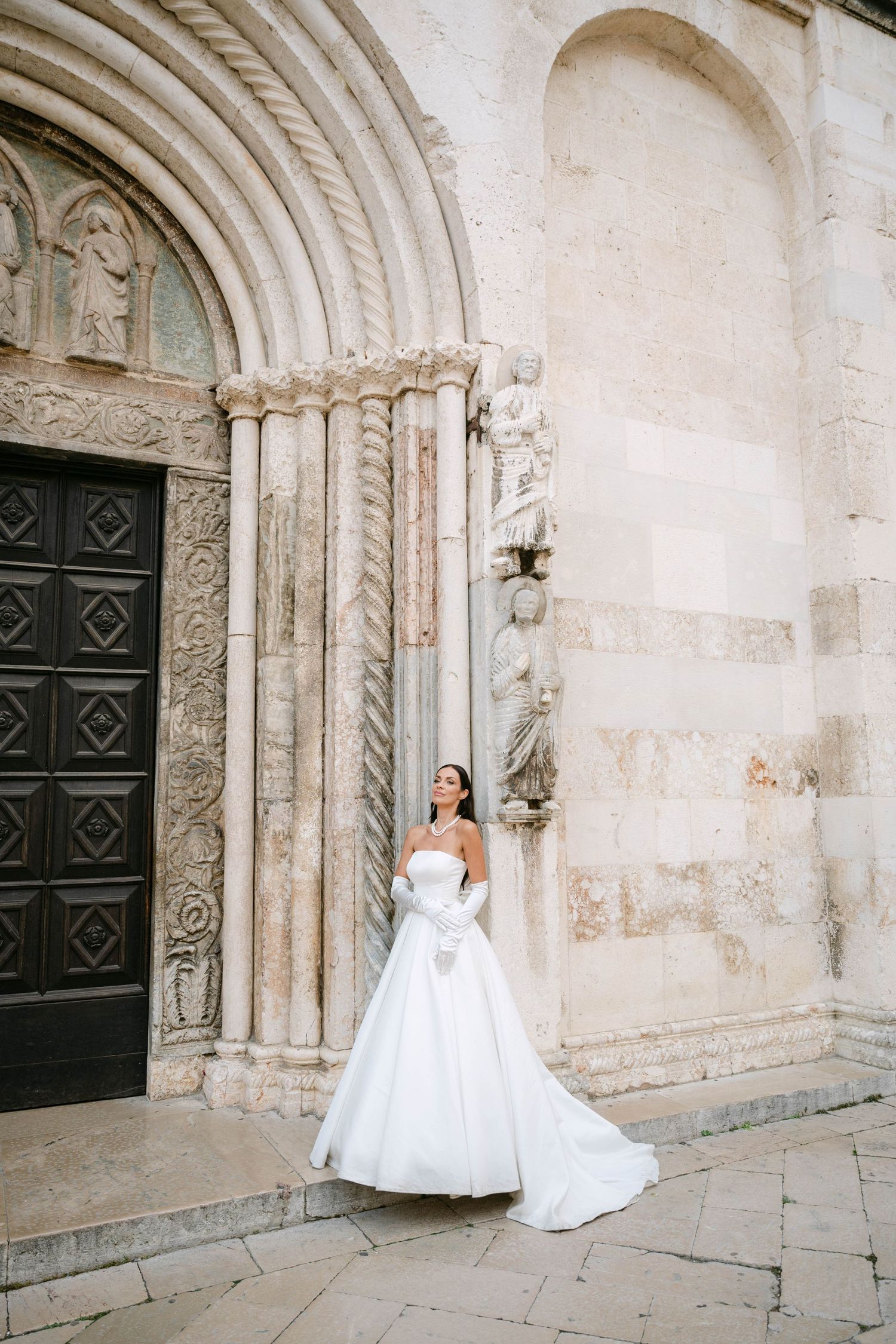 A bride in a flowing white gown poses in front of an ornate stone church entrance with decorative archways.