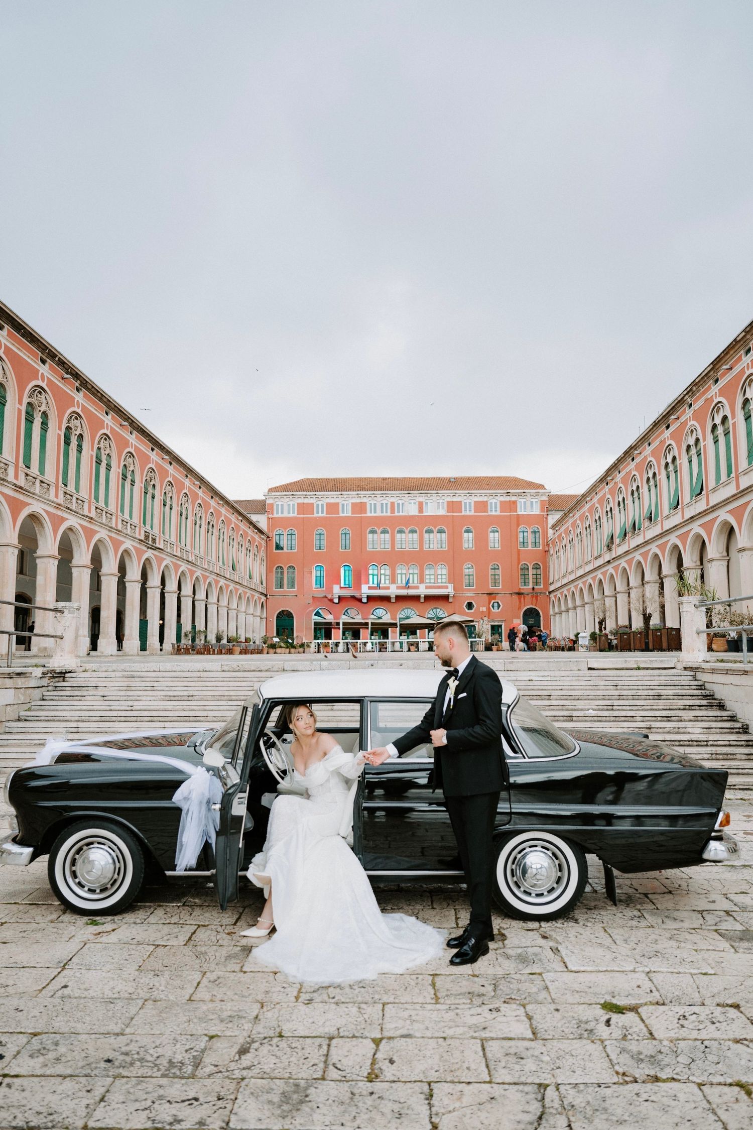 Bride and groom pose beside a vintage black car in a grand European square with red arched buildings.