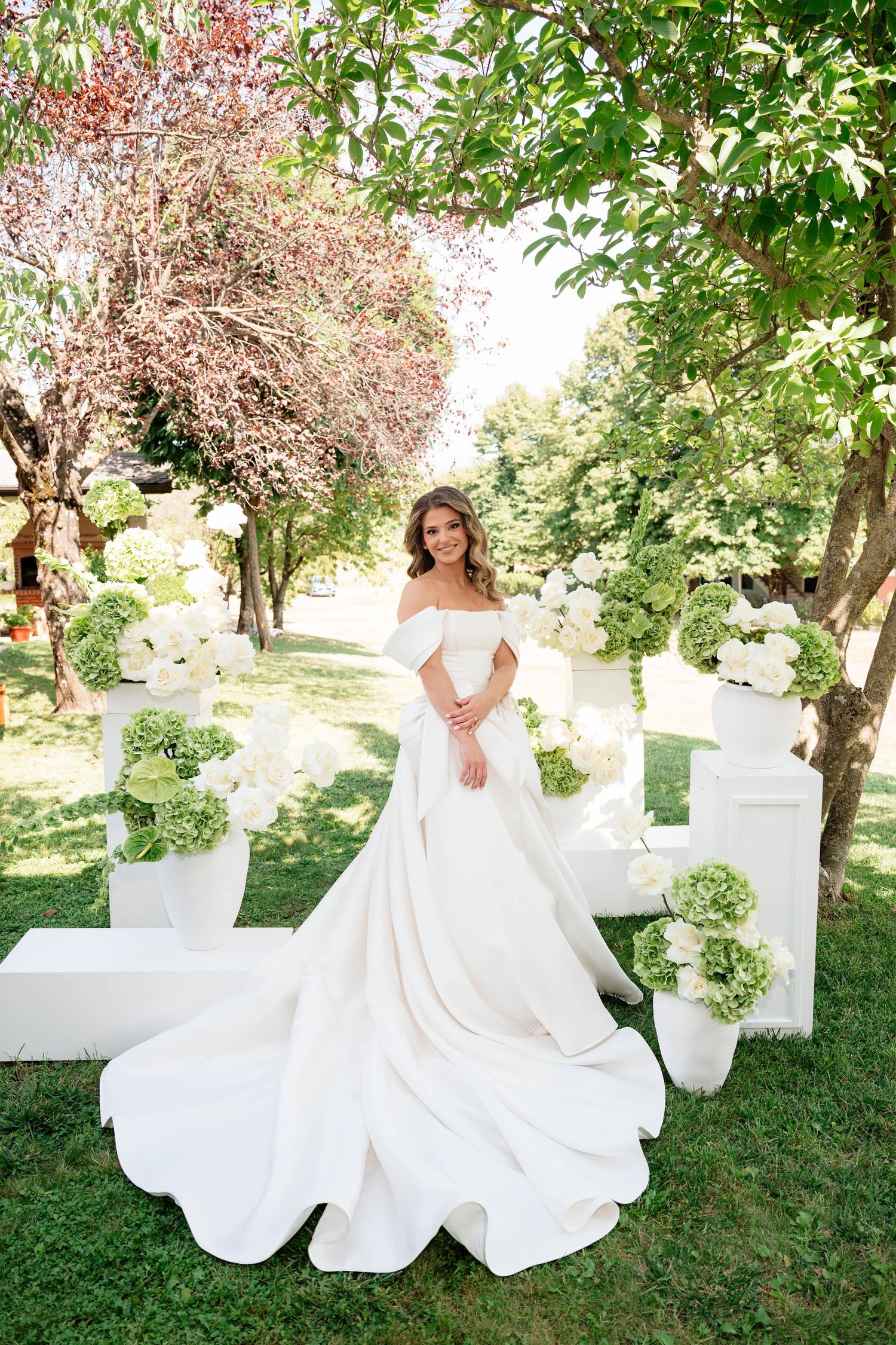 Bride in elegant off-shoulder white ball gown with long train poses outdoors among white floral arrangements and green trees.