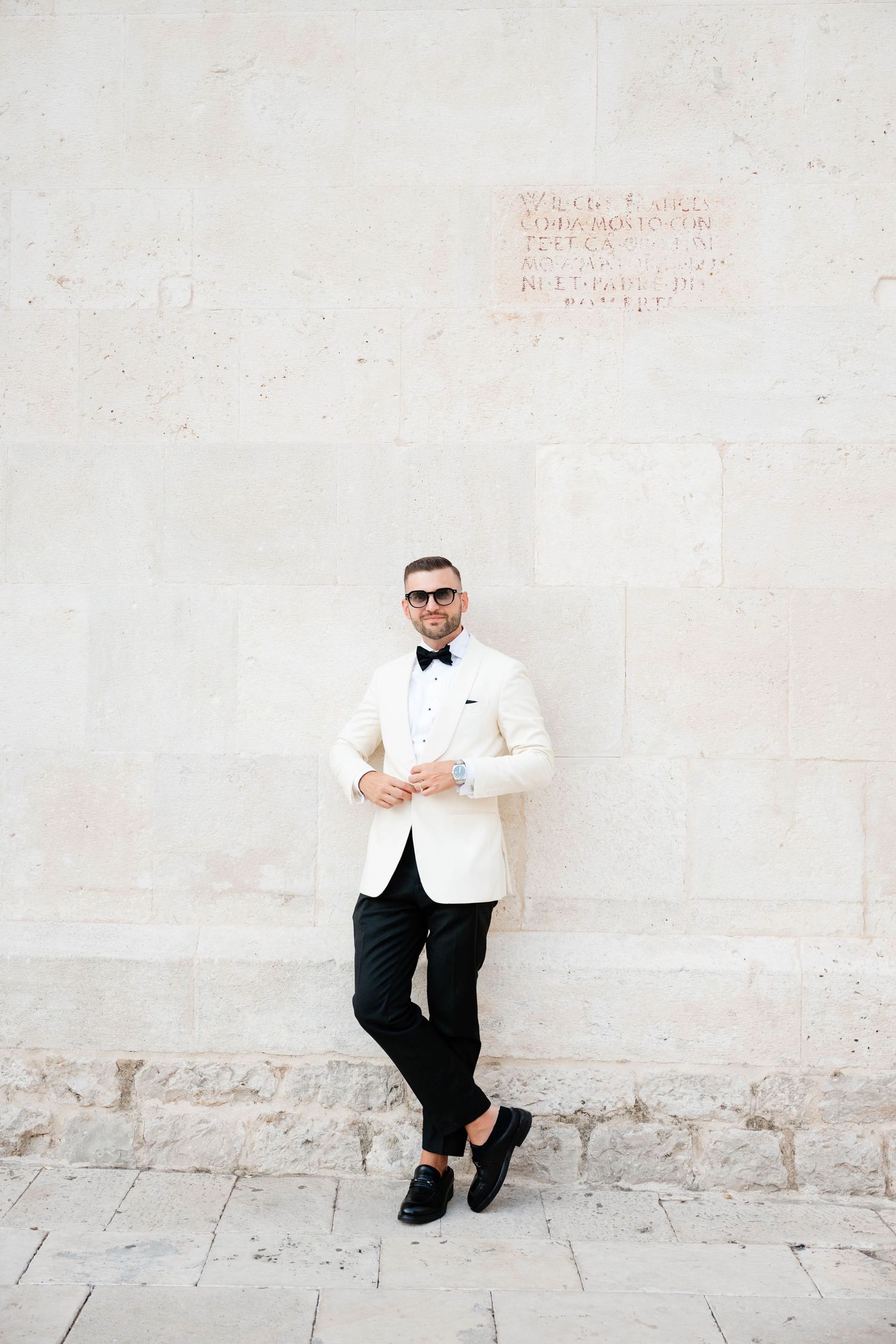 Stylish man in white tuxedo jacket, black bow tie, sunglasses, posing against a stone wall.