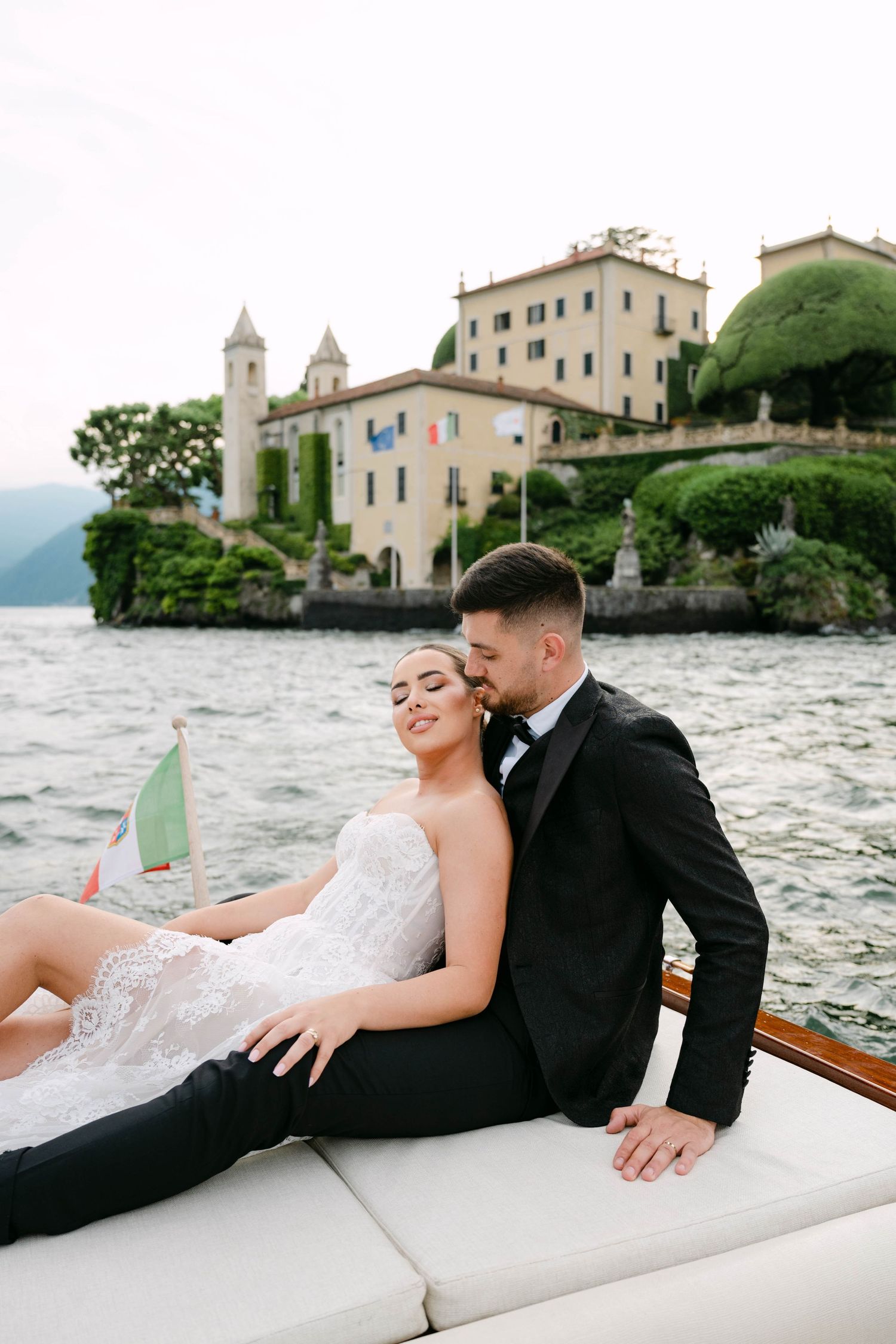 Bride and groom sitting on a luxury boat at Lake Como, Italy, with a historic villa and lush greenery in the background.