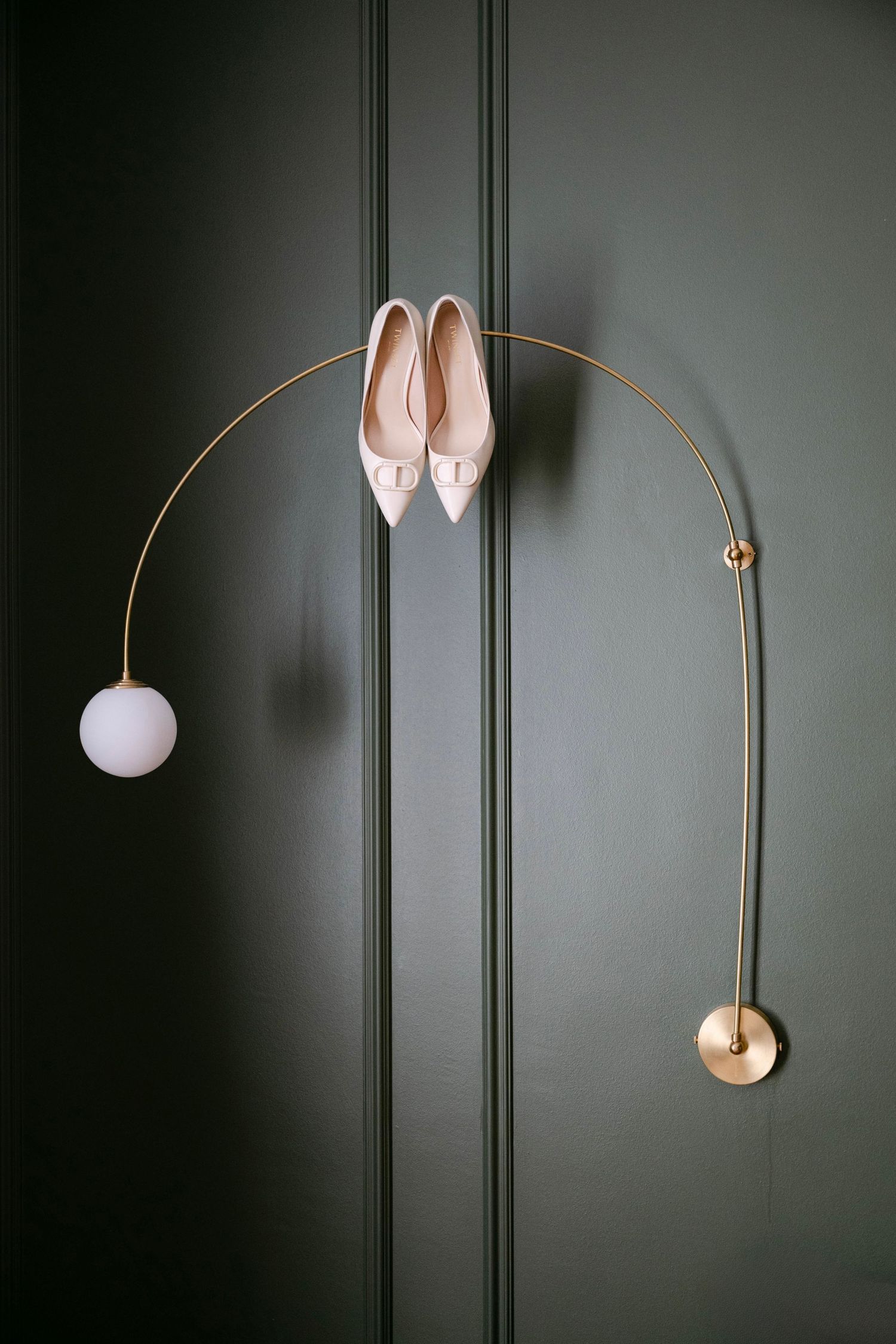White pointed ballet flats elegantly displayed on a curved gold wall lamp against a dark green wall.