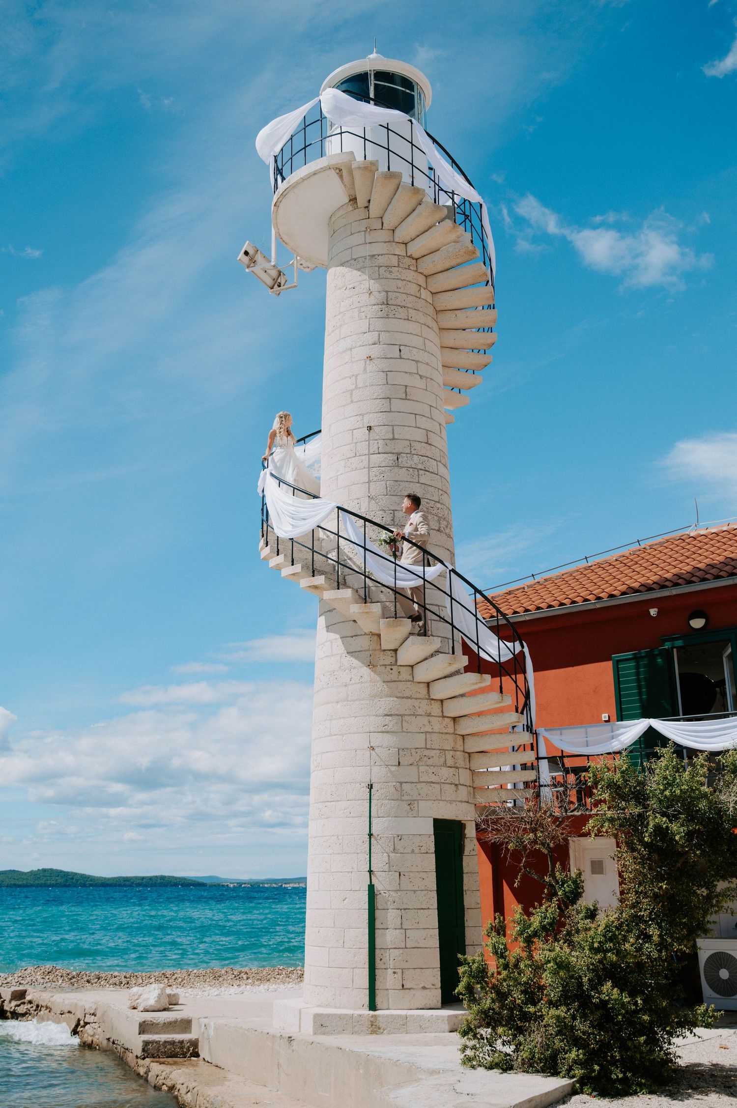 Spiral lighthouse staircase overlooking blue Mediterranean waters and red building.