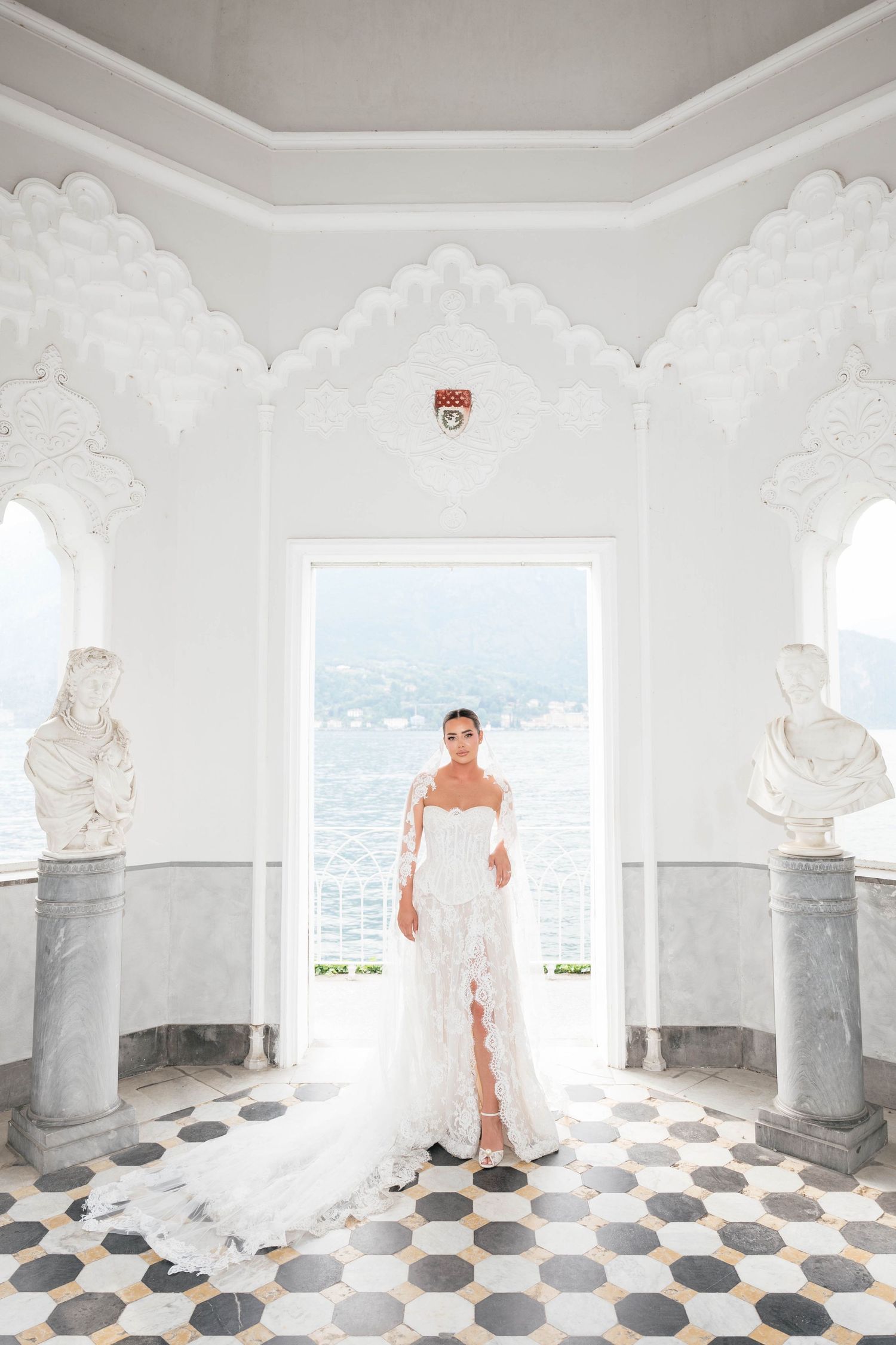 Bride in white lace gown stands in elegant marble villa with arched windows overlooking a scenic lake.