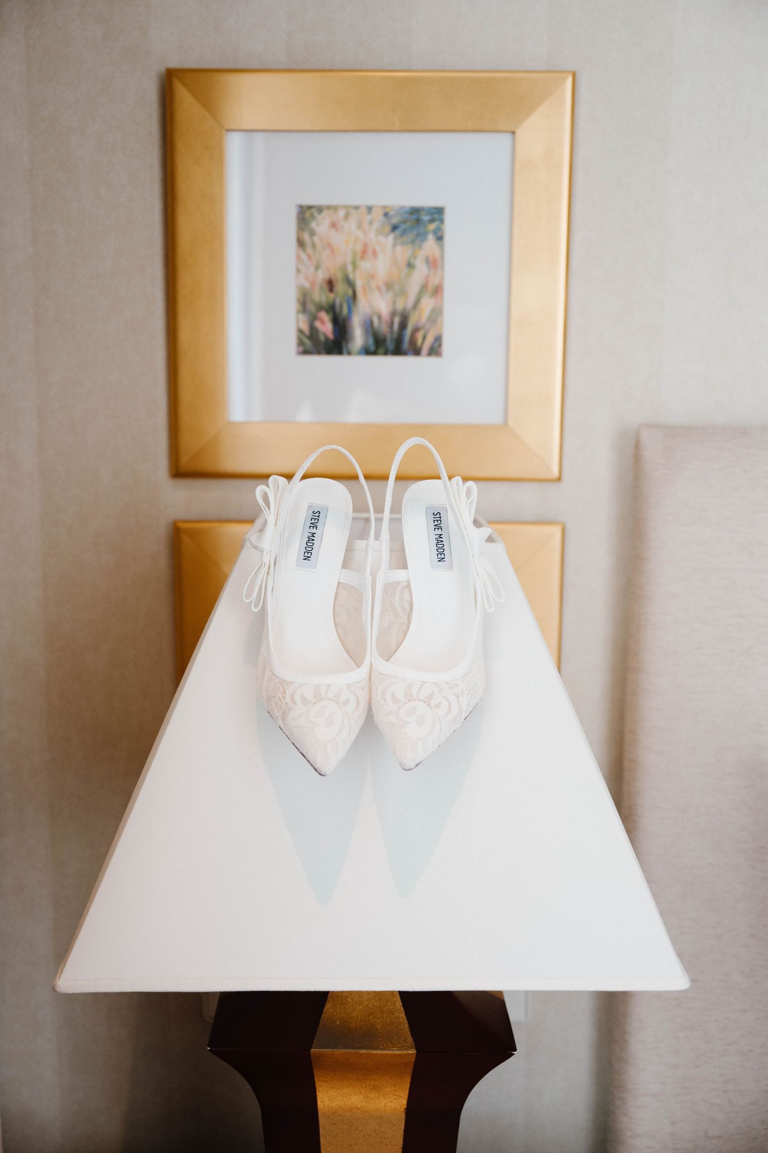 Steve Madden white lace slingback wedding heels displayed on a white table with a gold-framed floral artwork in the background.