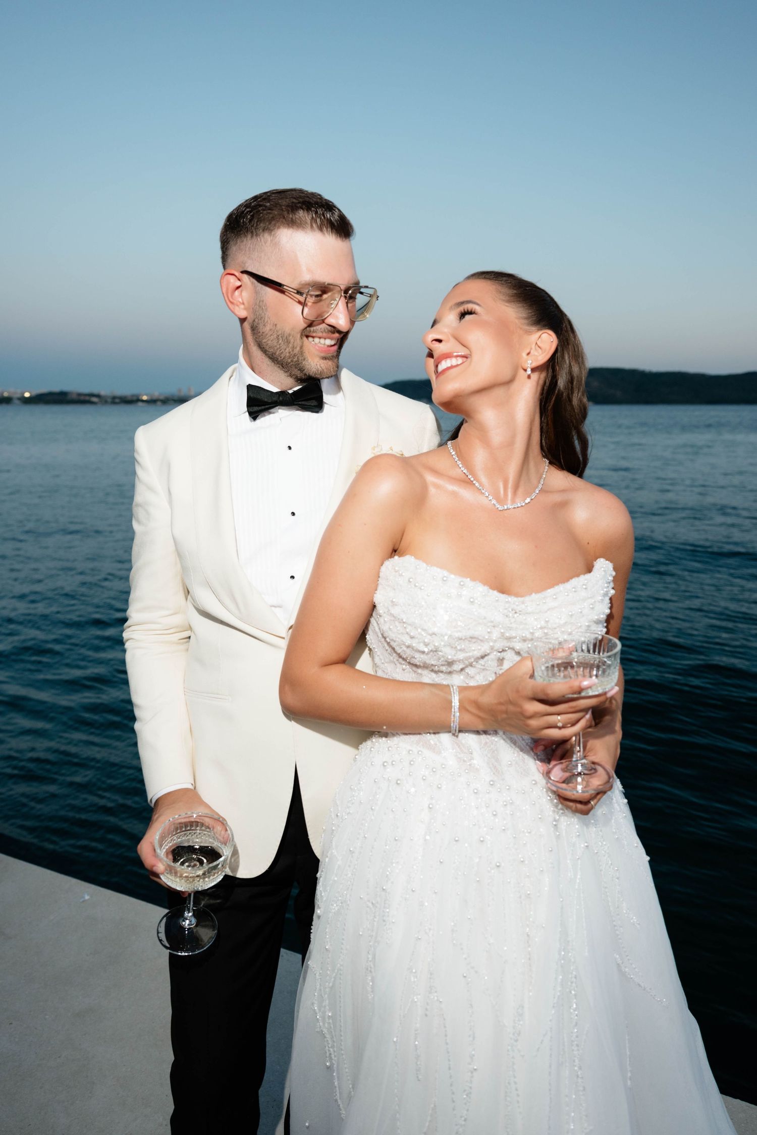 Bride and groom holding champagne glasses by the sea at sunset, wearing white wedding attire.