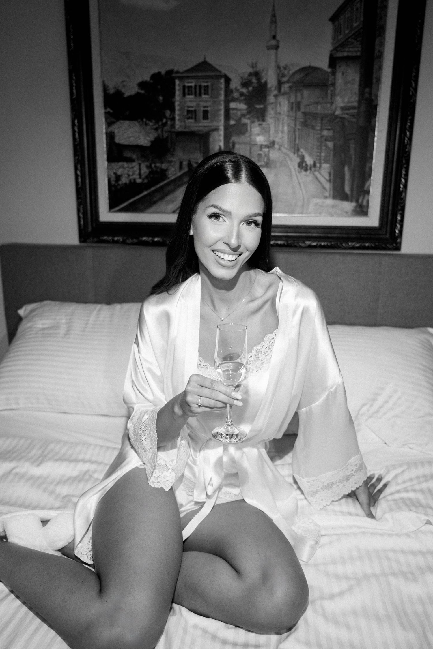 Black and white photo of someone in a white silk robe sitting on a bed with a drink, with framed artwork on wall.