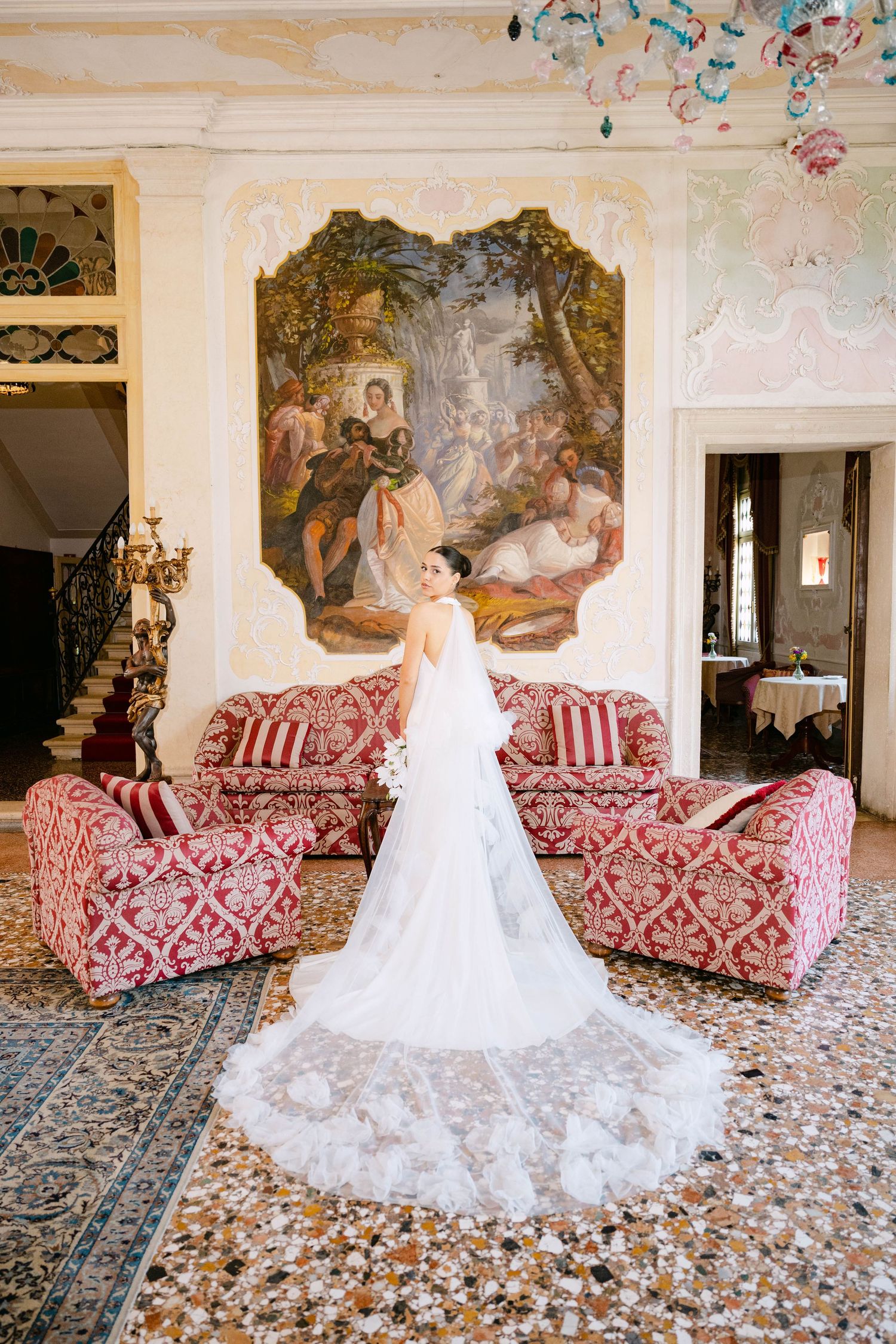 Elegant white wedding gown with long train displayed in luxurious room with vintage furniture and ornate wall mural.
