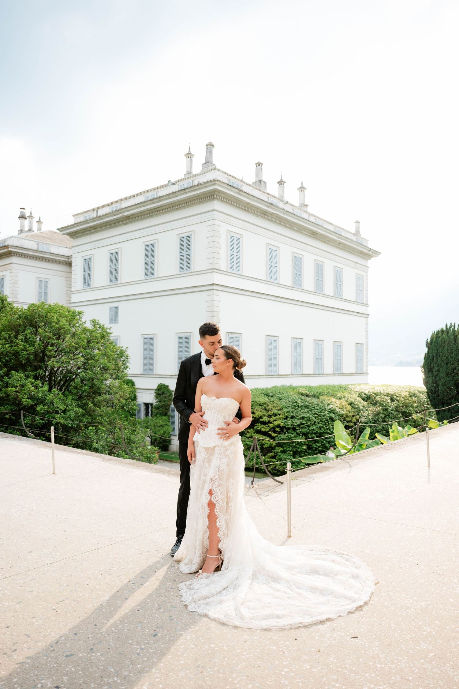 Bride and groom embrace before a grand white villa on Lake Como, Italy, in an elegant lace wedding gown and black tuxedo.