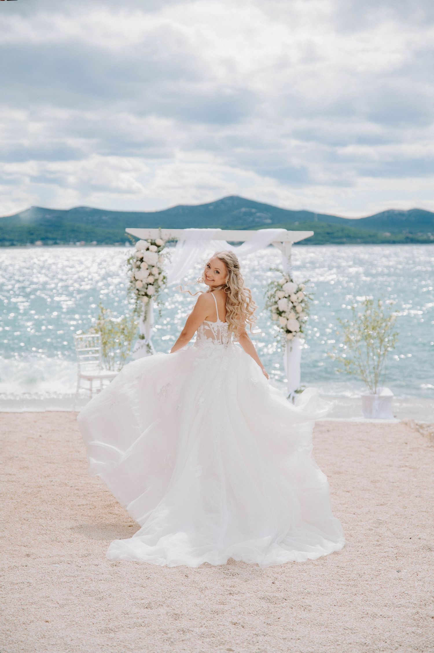 A bride in a flowing gown spins on a beach with mountains and an ocean backdrop.