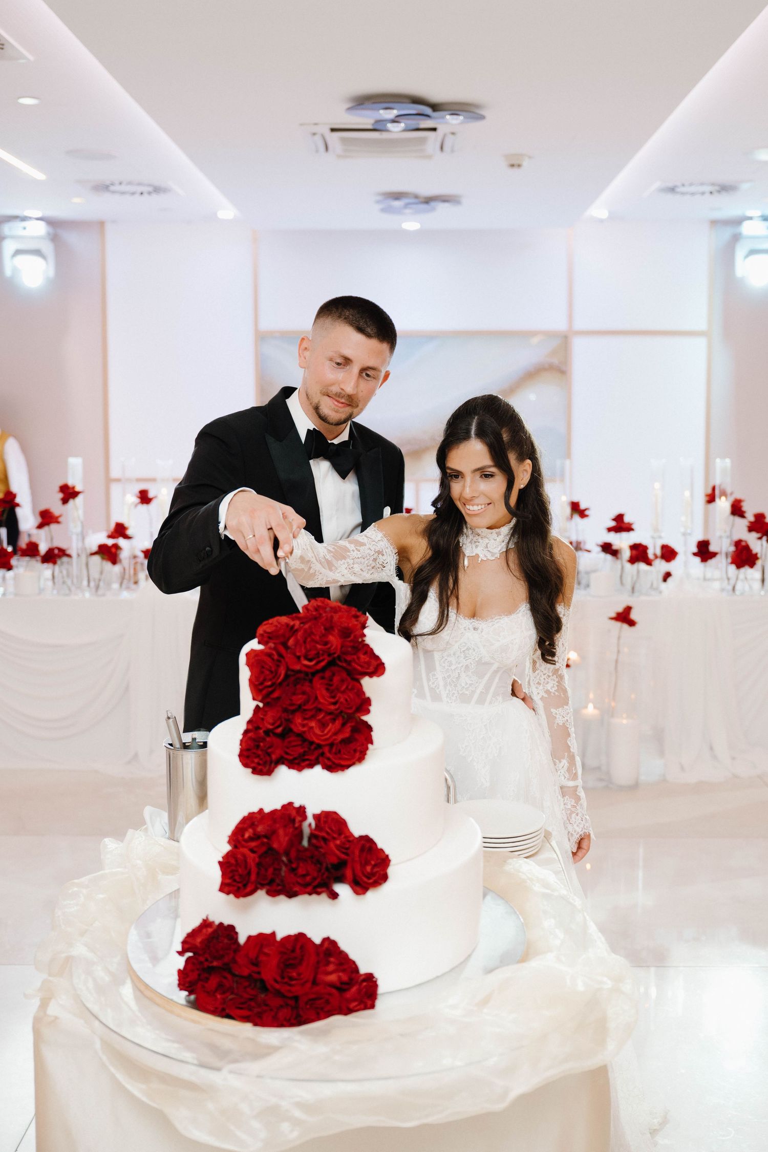 Bride and groom cutting a white tiered wedding cake adorned with red roses in an elegant white reception hall.